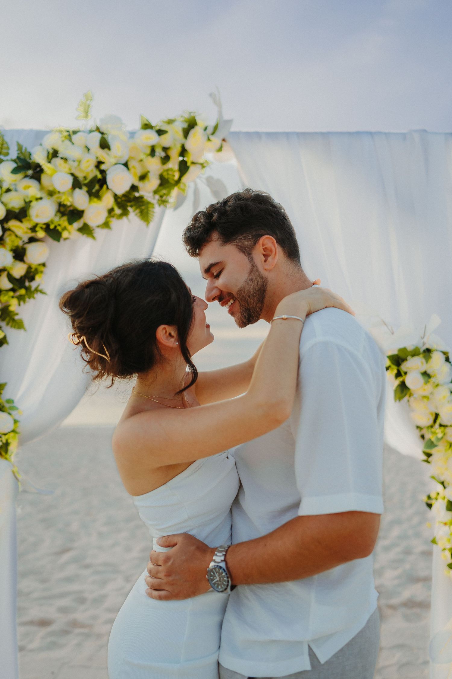 Happy couple embraces and smiles under floral arch decorated with white roses on sandy beach.