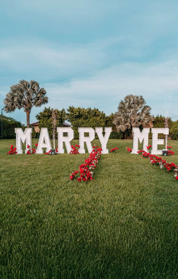 Large illuminated MARRY ME letters with red roses on green grass surrounded by palm trees.