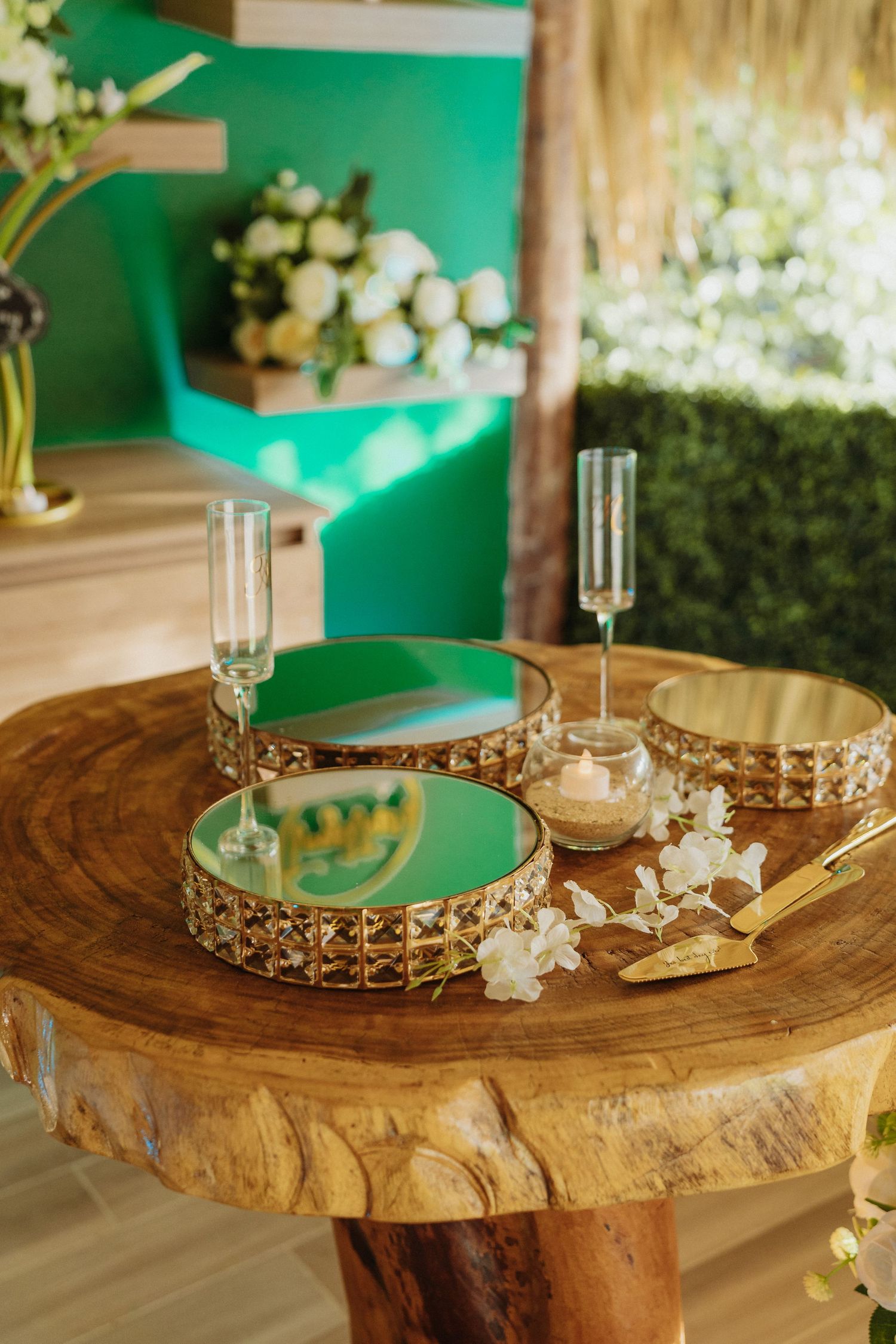 Rustic wooden slab table with gold-rimmed green trays, champagne flutes, and candles at tropical wedding venue.
