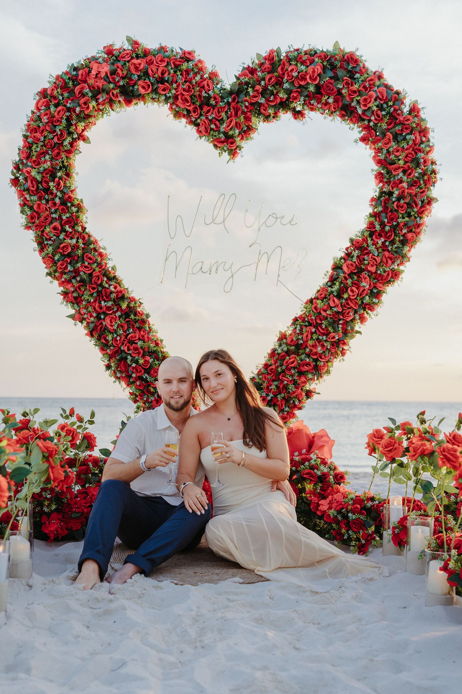 Couple sitting beneath a red floral heart arch with Will You Marry Me sign on a beach.