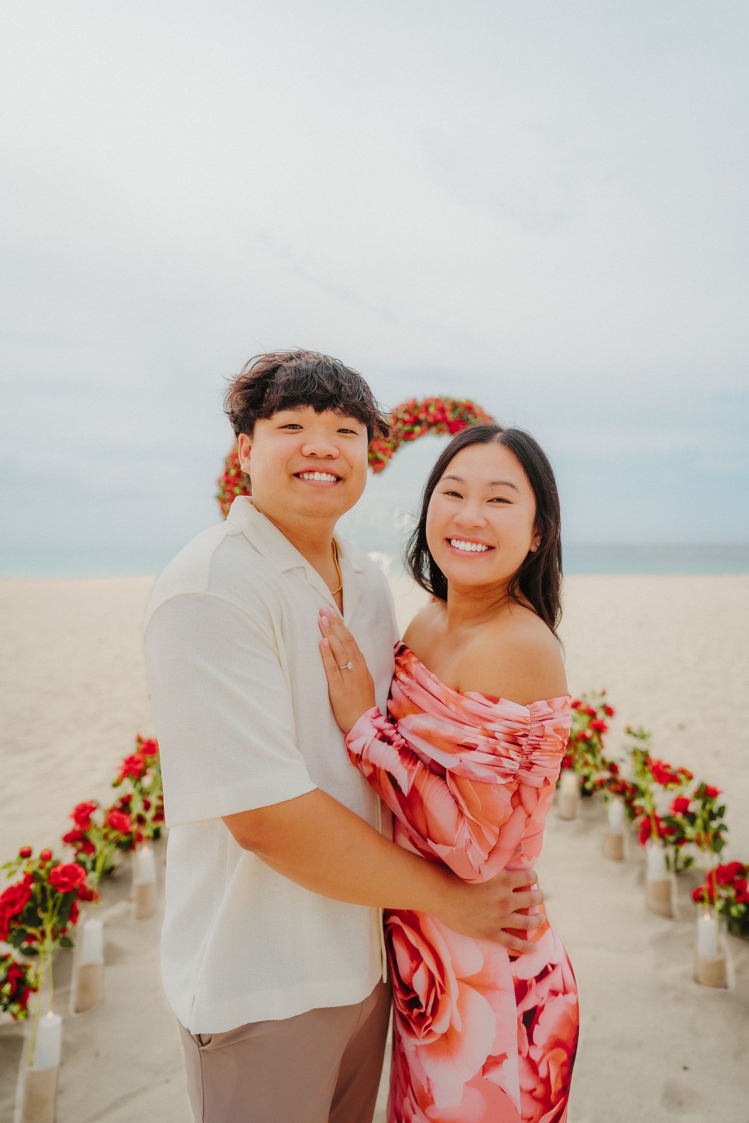 Happy couple embracing and smiling at a romantic red rose beach proposal setup.