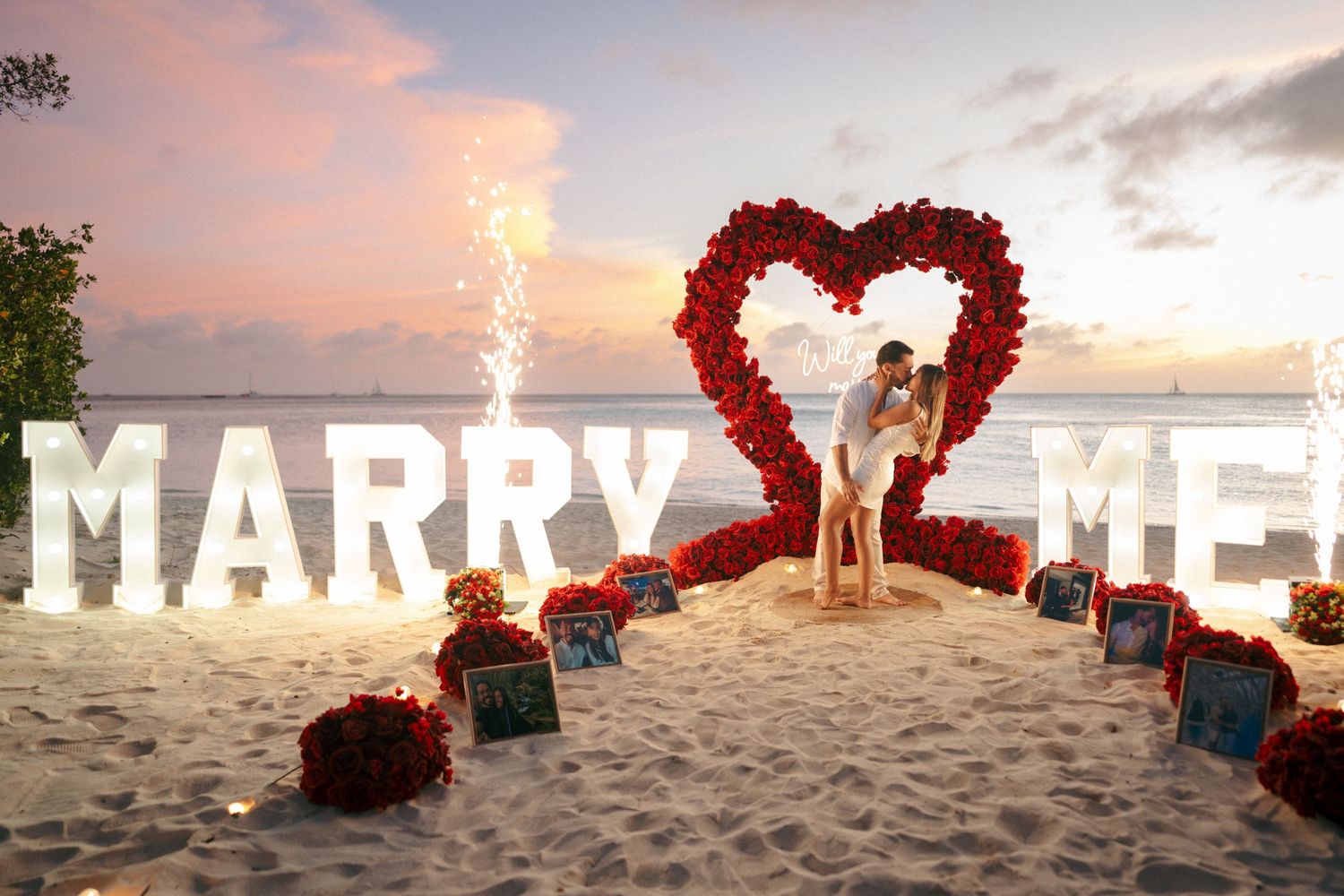 Couple embracing inside a red rose heart under glowing MARRY ME letters on a beach at sunset, a romantic proposal setup.
