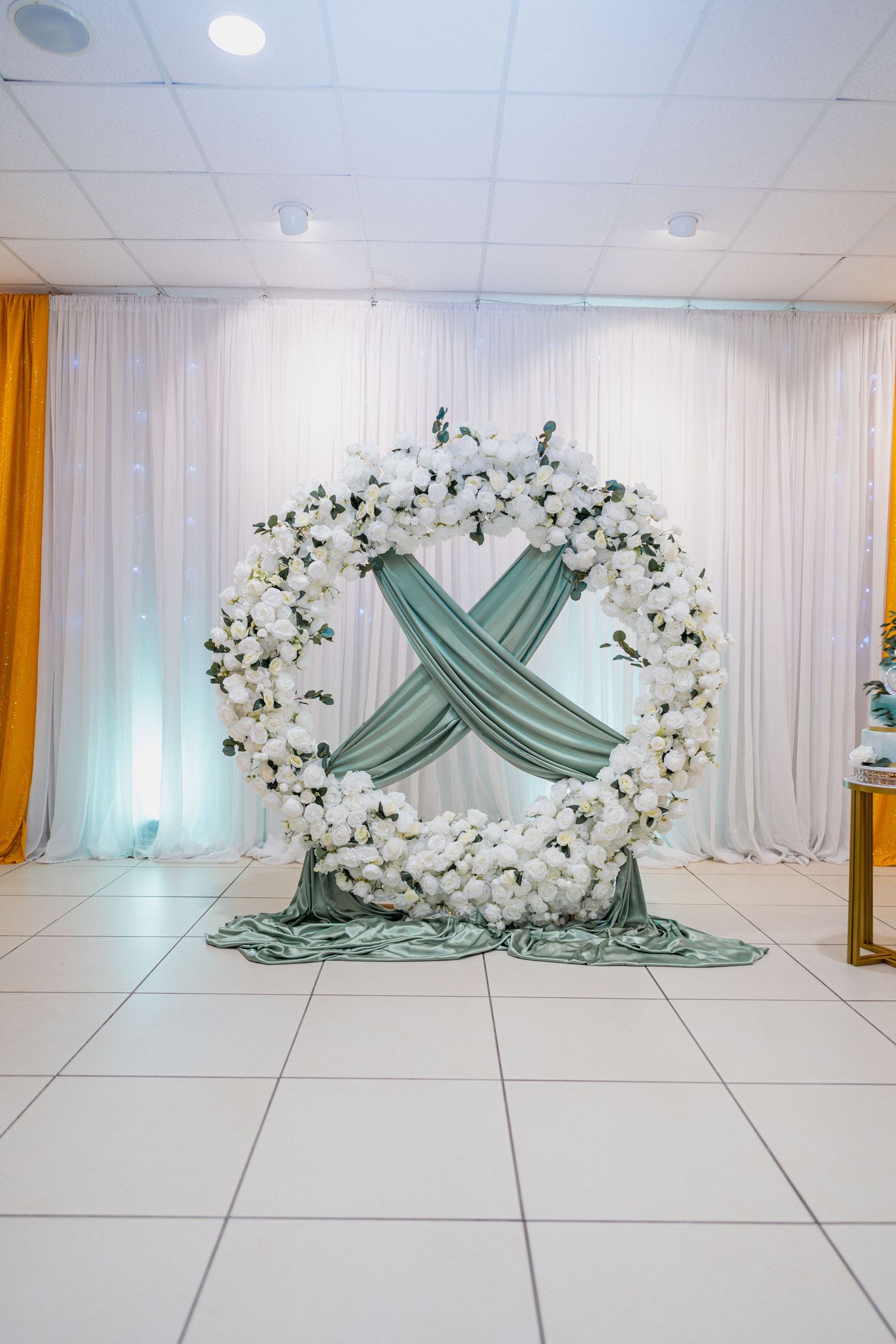 Elegant white floral double-ring wedding arch with sage green draping in decorated event hall.