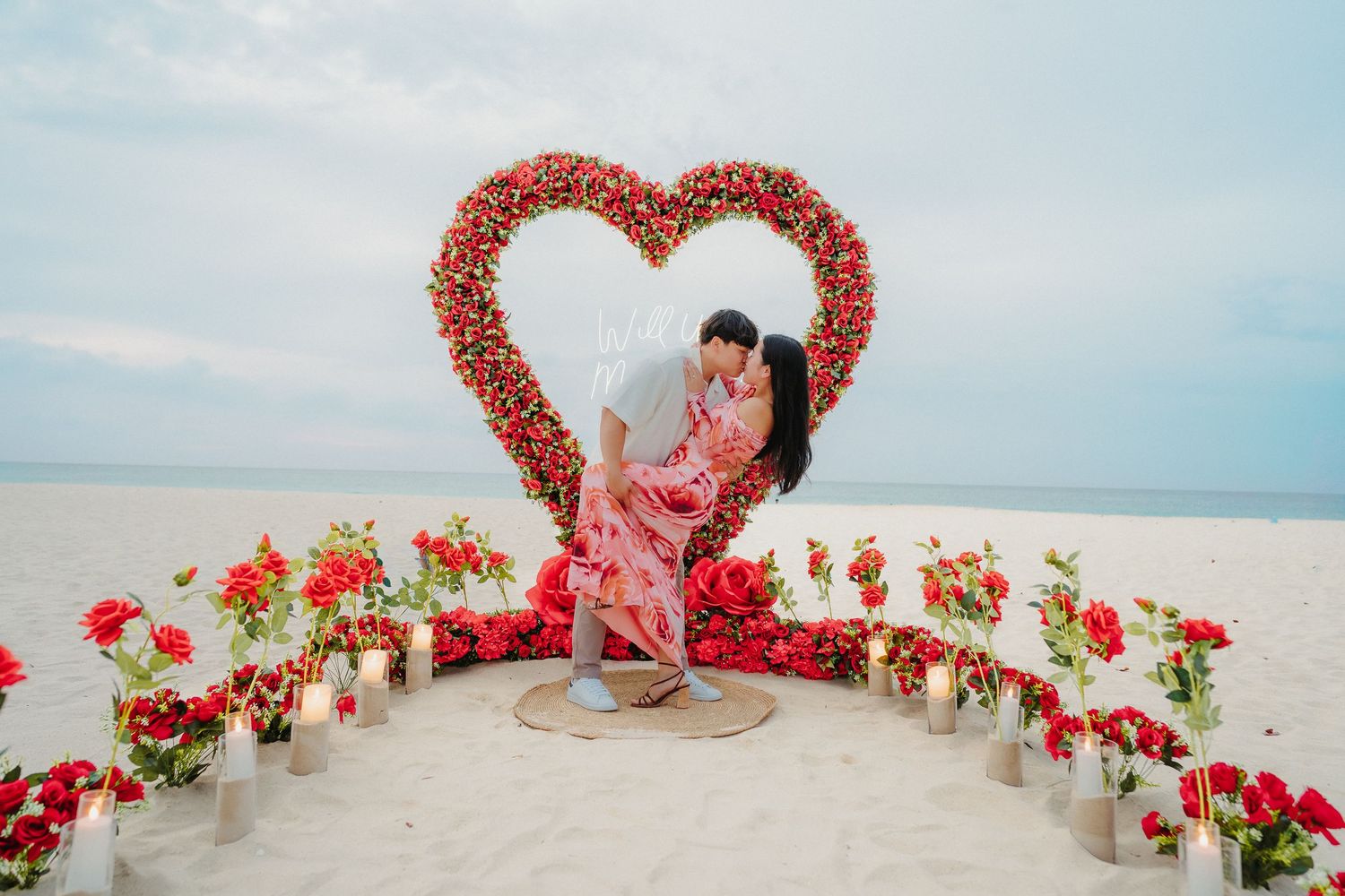 Couple sharing a romantic kiss in front of a stunning red floral heart arch on the beach.