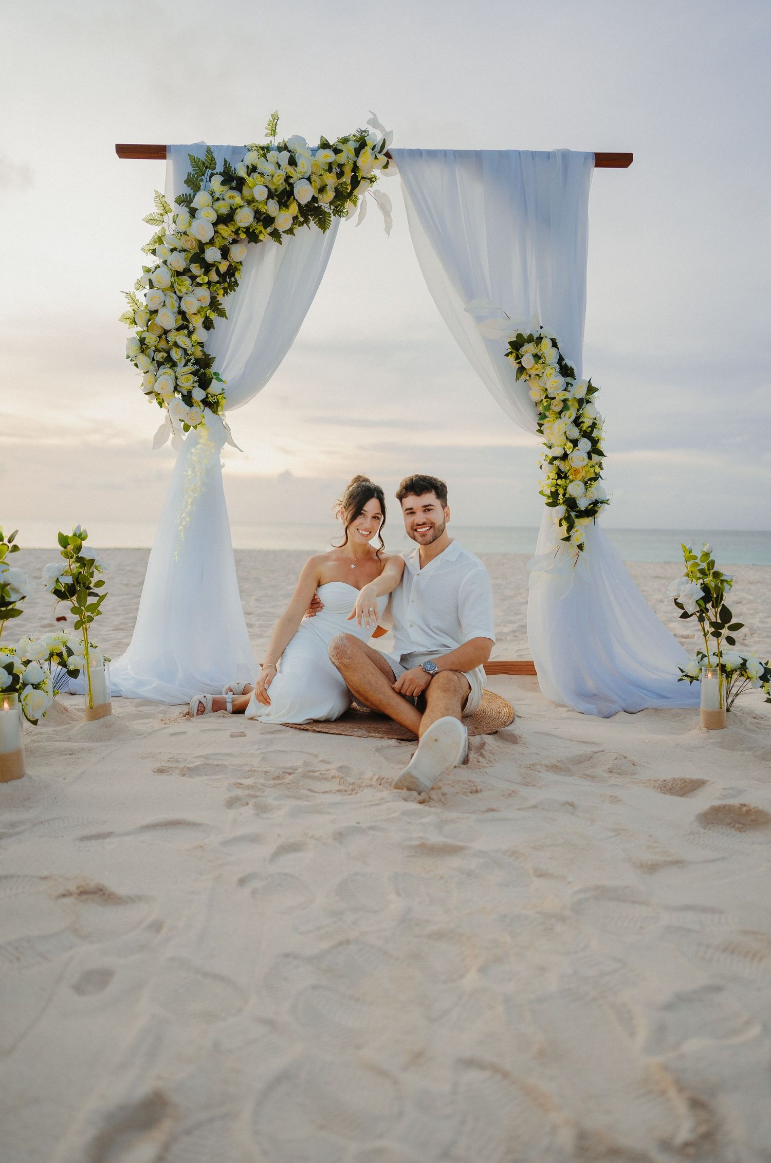 Couple sitting together on sandy beach under white floral wedding arch at sunset.