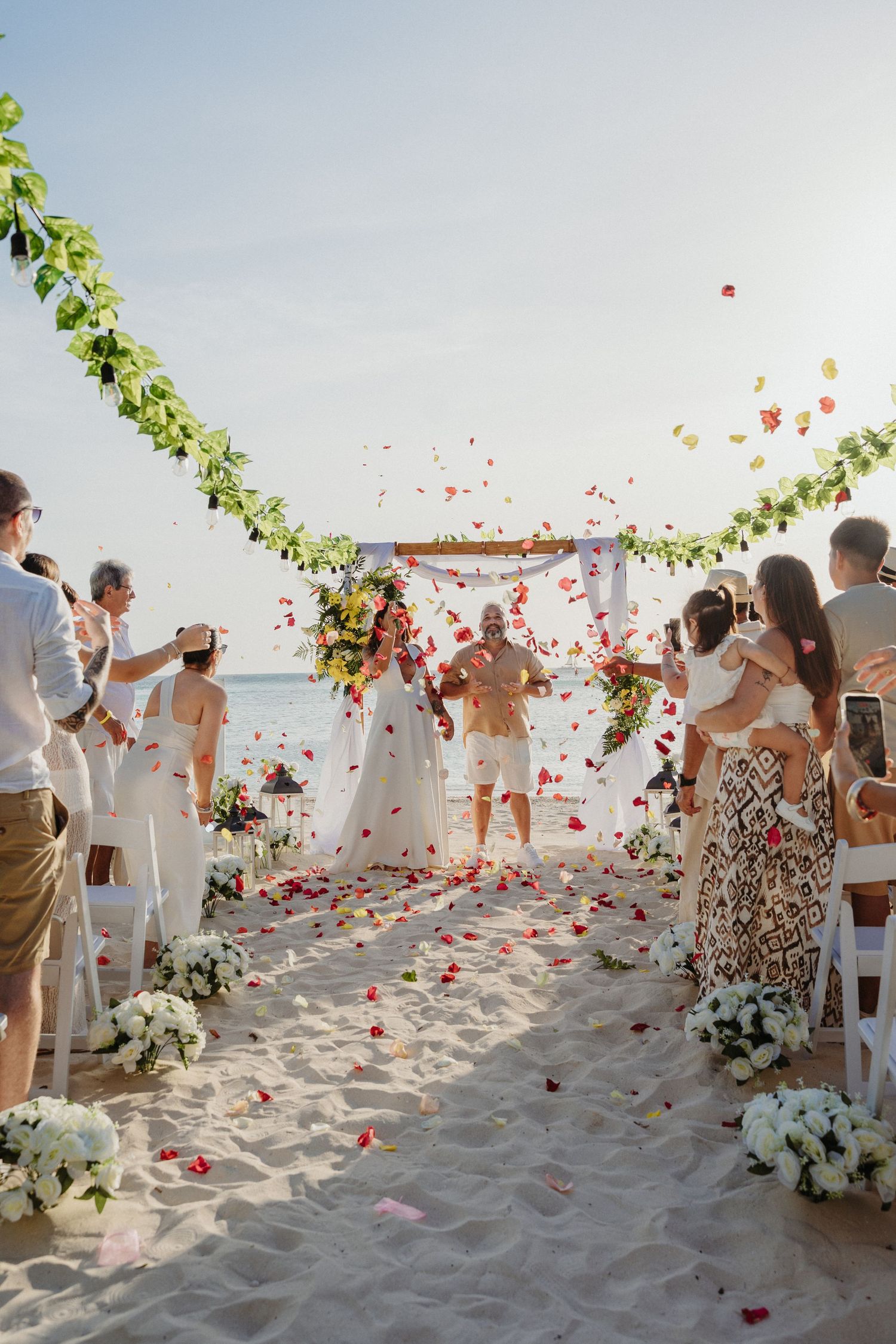 Joyful beach wedding ceremony with guests tossing colorful flower petals as couple walks down the aisle.