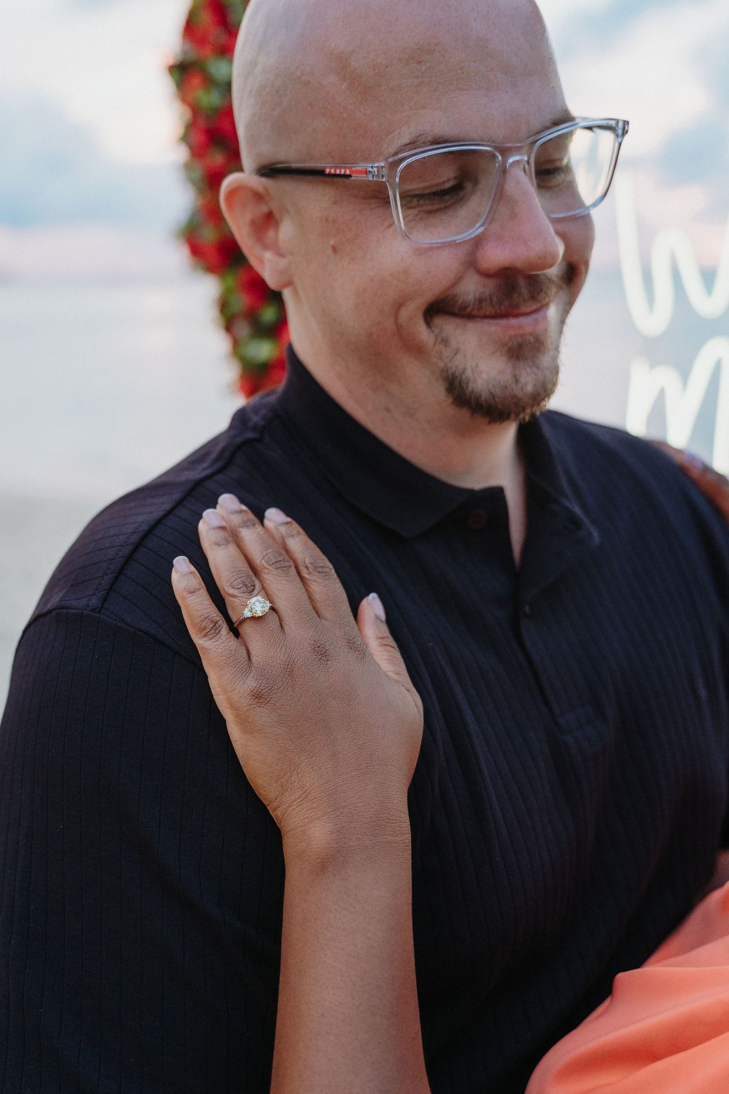 Newly engaged woman rests hand with diamond ring on fiances chest during beach proposal.