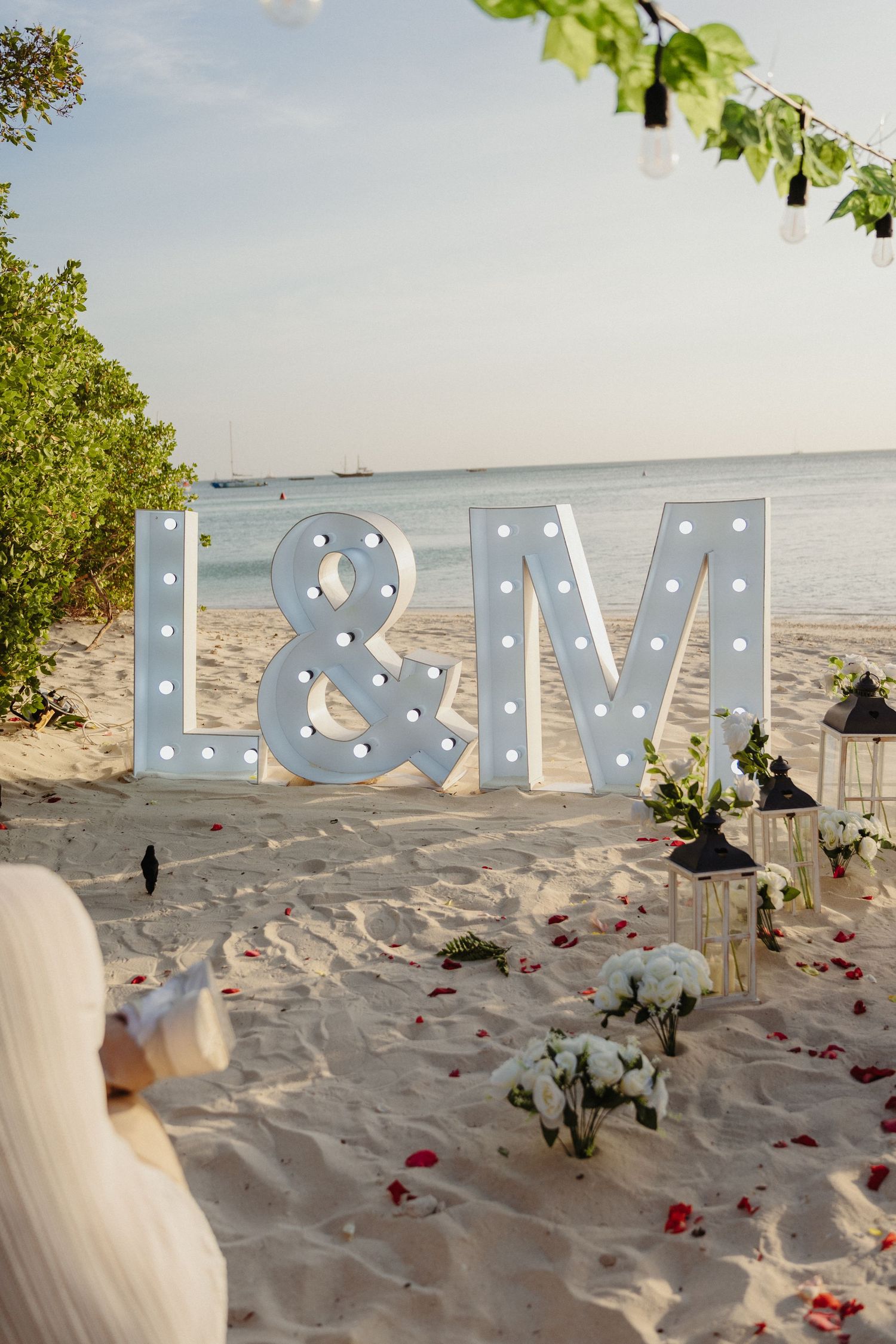 Beach wedding decor with marquee letters L&M, white lanterns, roses, and red petals on sandy shore.