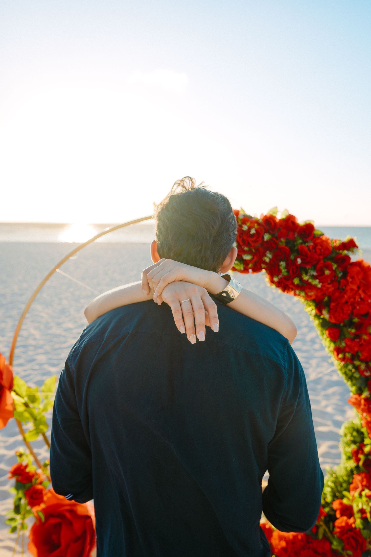 Person being embraced from behind near red floral arch on sunny beach, showing ring on hand.