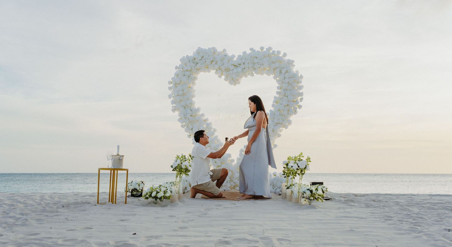 Man proposes to woman on beach with white floral heart arch backdrop.