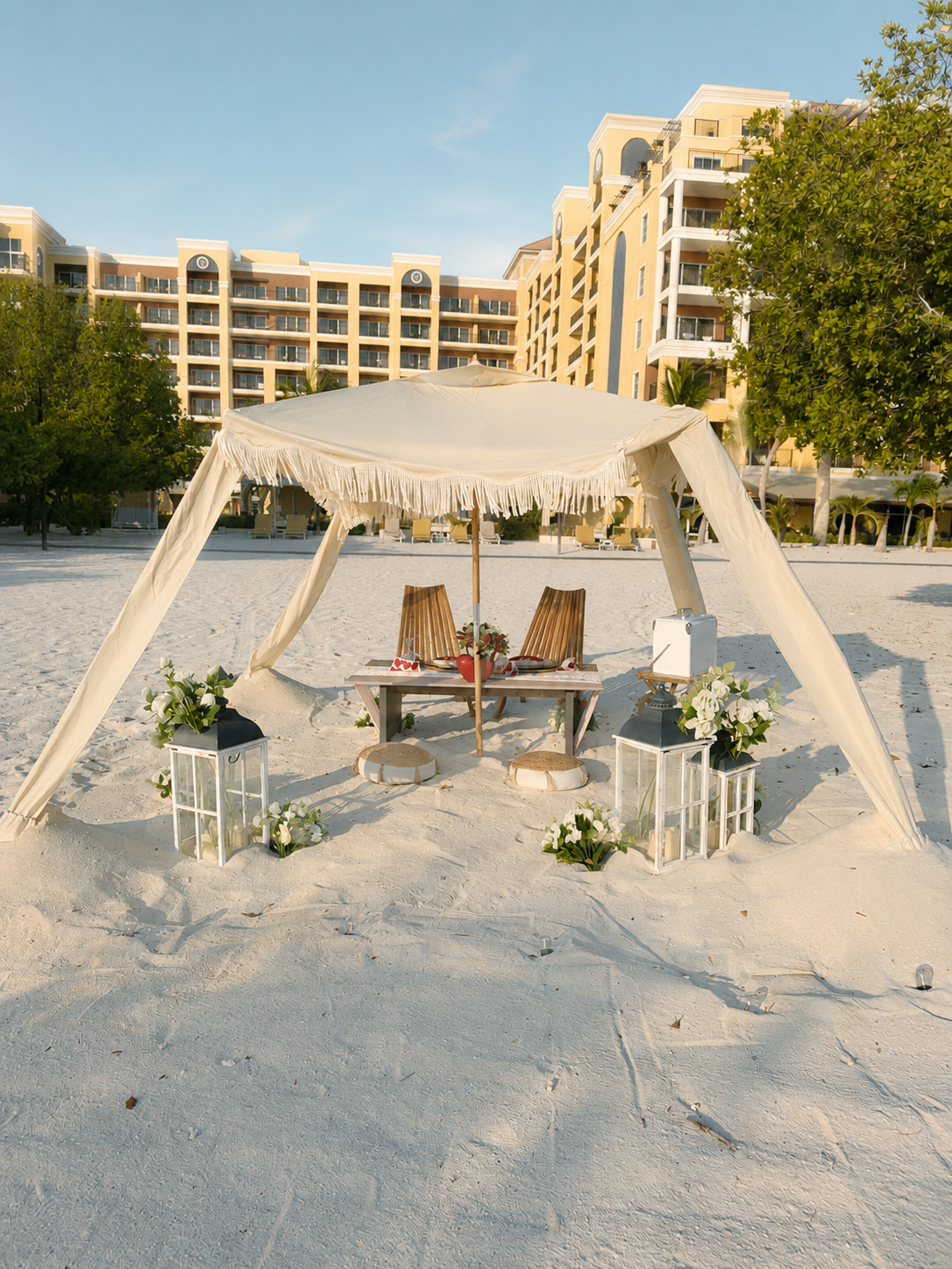 Elegant beach cabana with fringe canopy, floral arrangements, and lanterns set for a romantic occasion with resort backdrop.