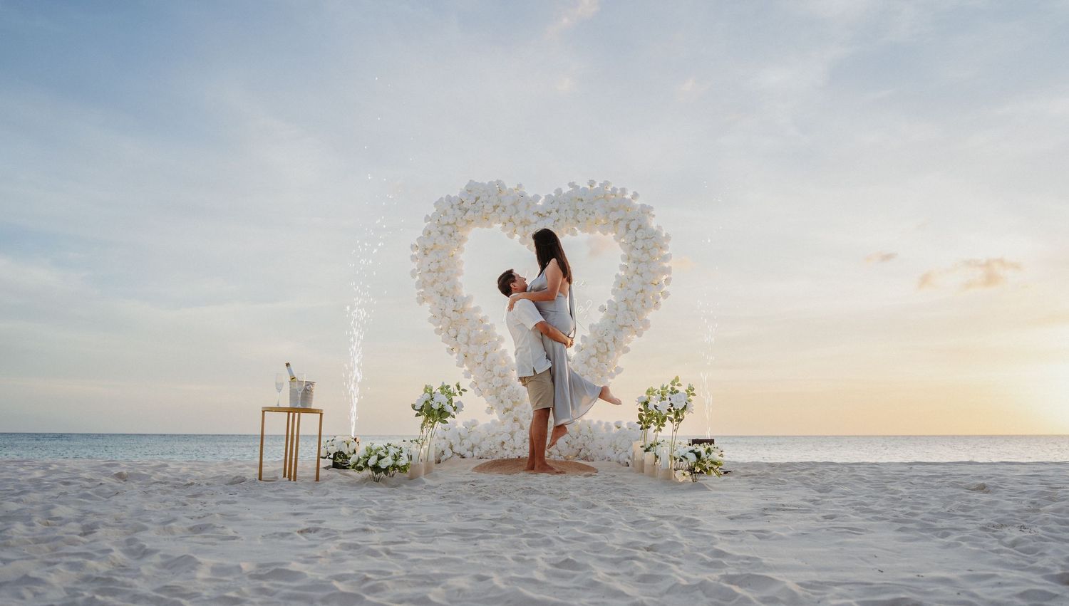 Couple embraces in front of white floral heart arch on a serene beach.