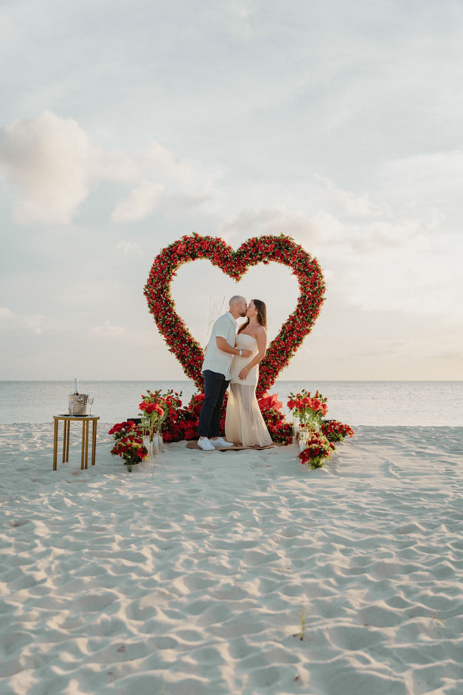 Couple shares a kiss under a red rose heart arch on a white sand beach.