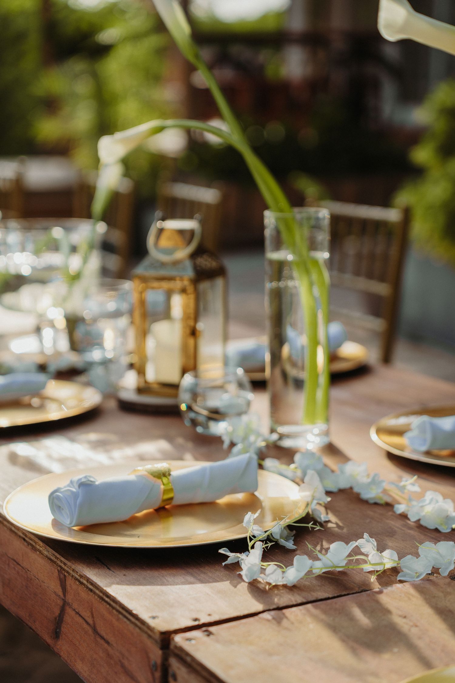 Gold wedding table setting with white napkins, candles, and floral centerpieces outdoors.
