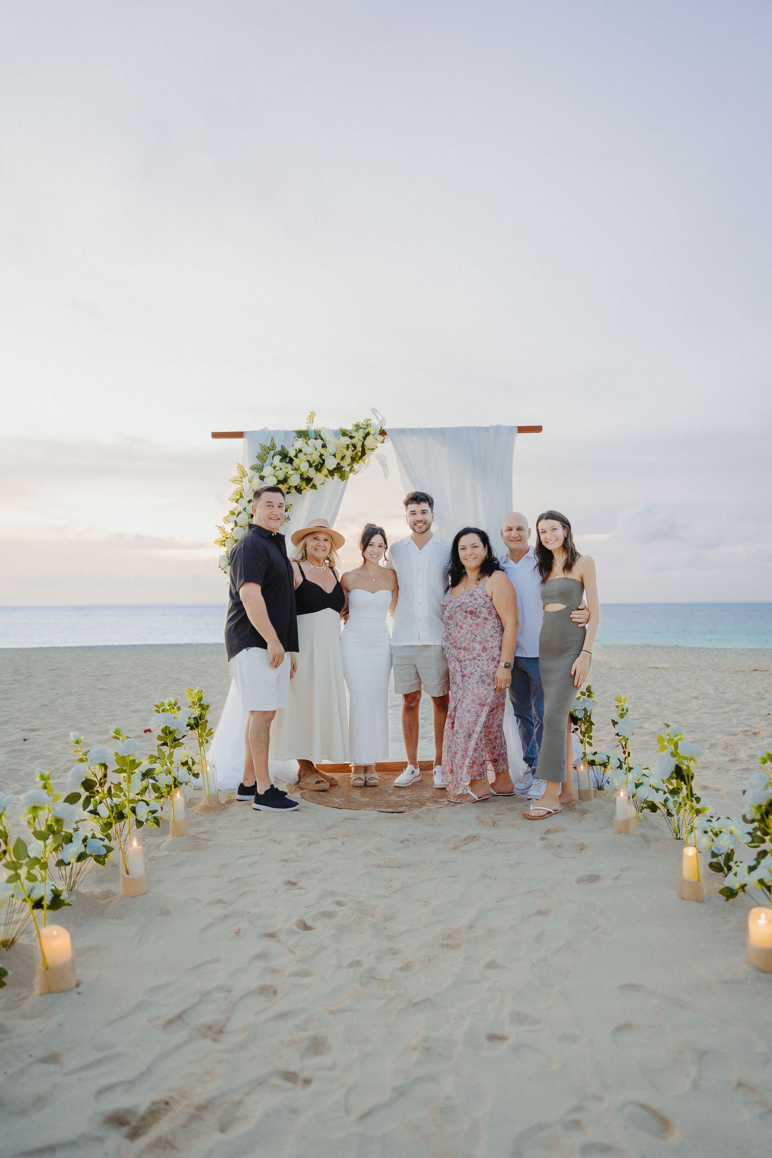 Wedding party poses together in front of white floral arch on sandy beach at sunset.