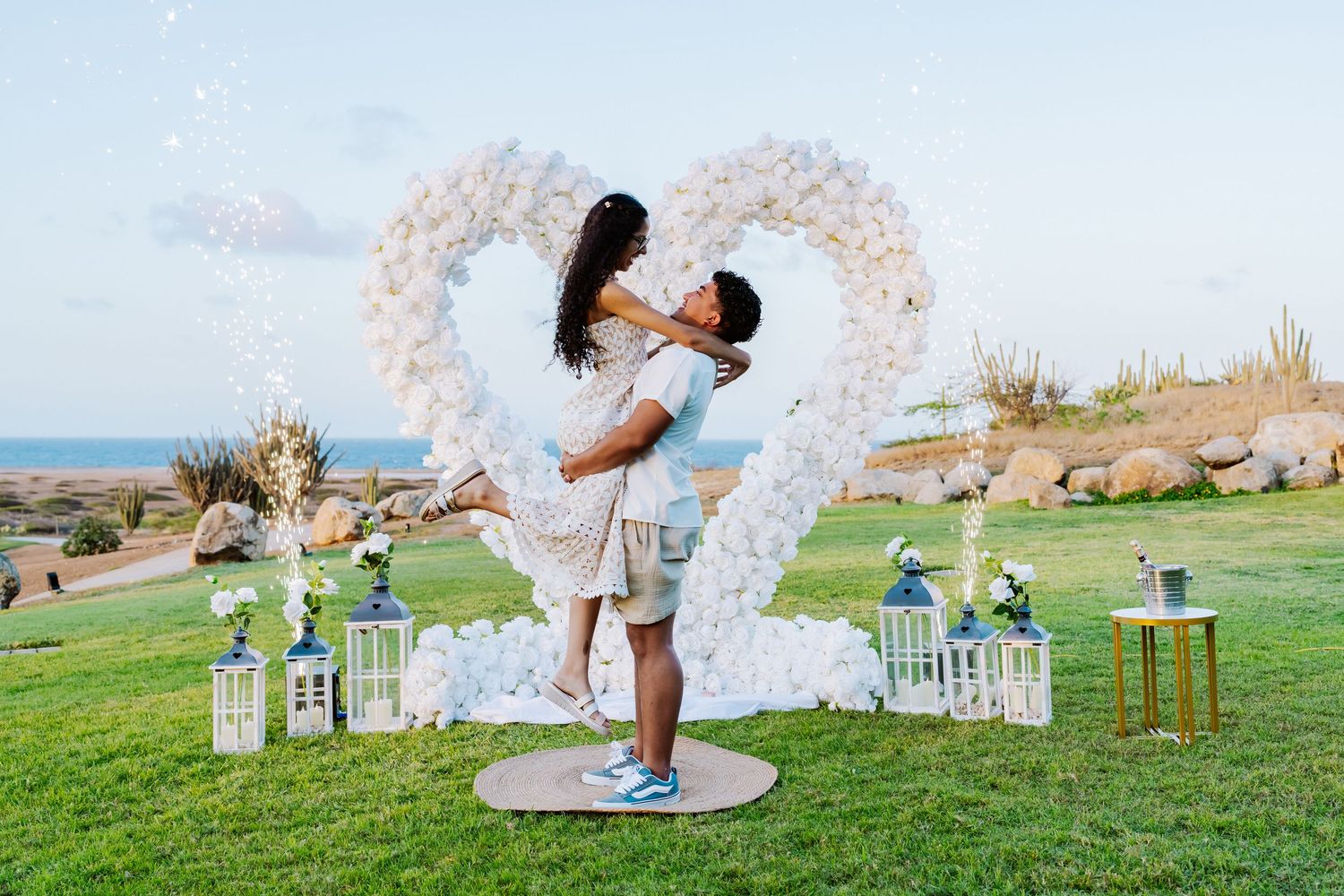 Joyful couple celebrating with man lifting woman in front of white floral heart arch outdoors.