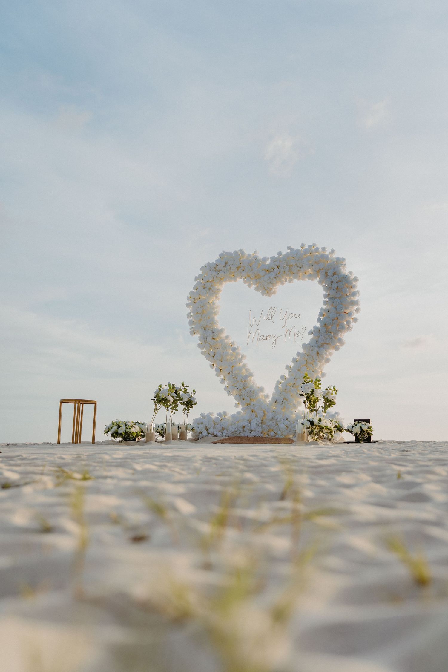 White floral heart-shaped wedding arch on a sandy beach with a cloudy sky backdrop.