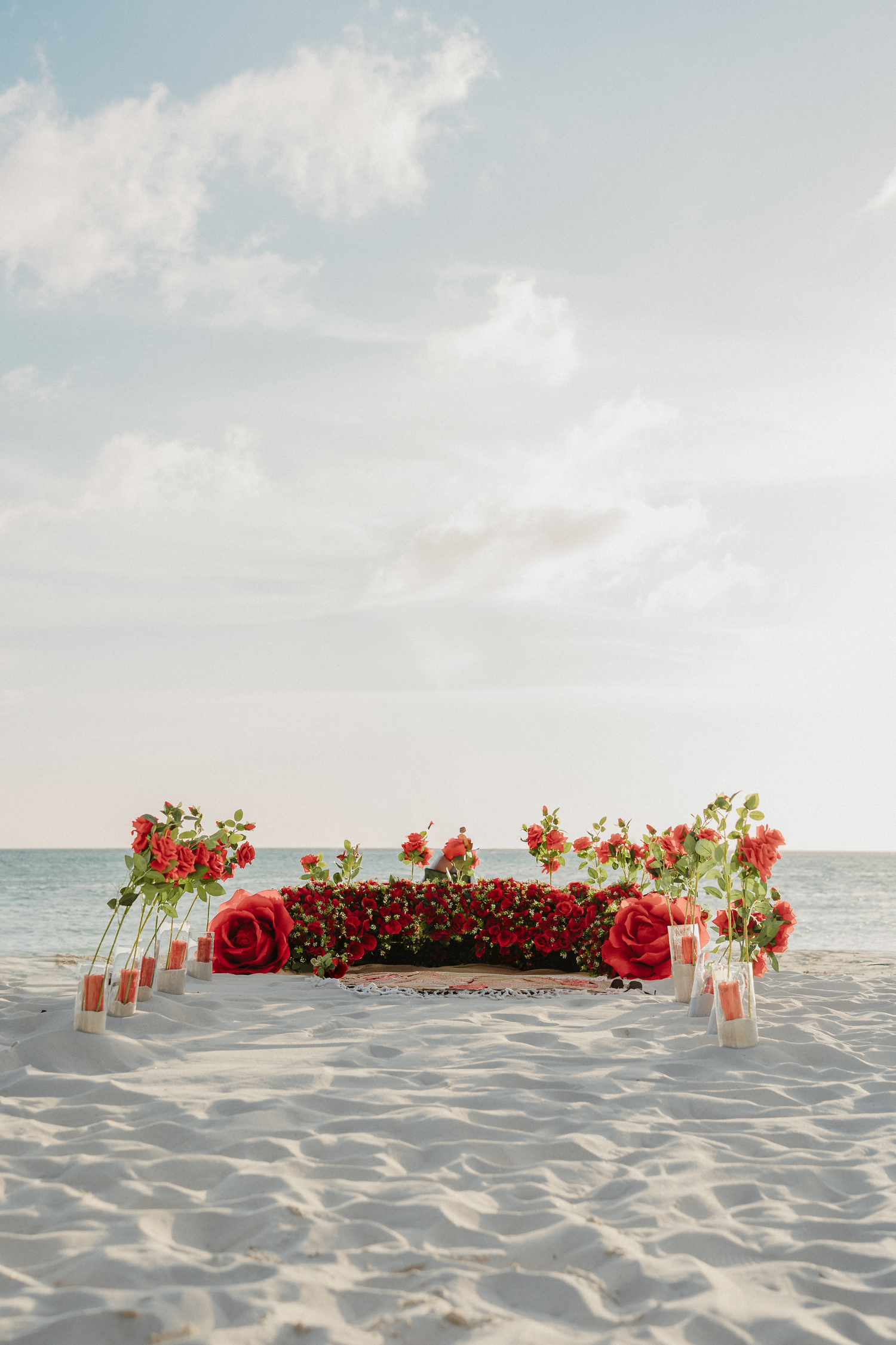 Romantic beach proposal setup with red roses, floral arch, and candles on white sand by the ocean.