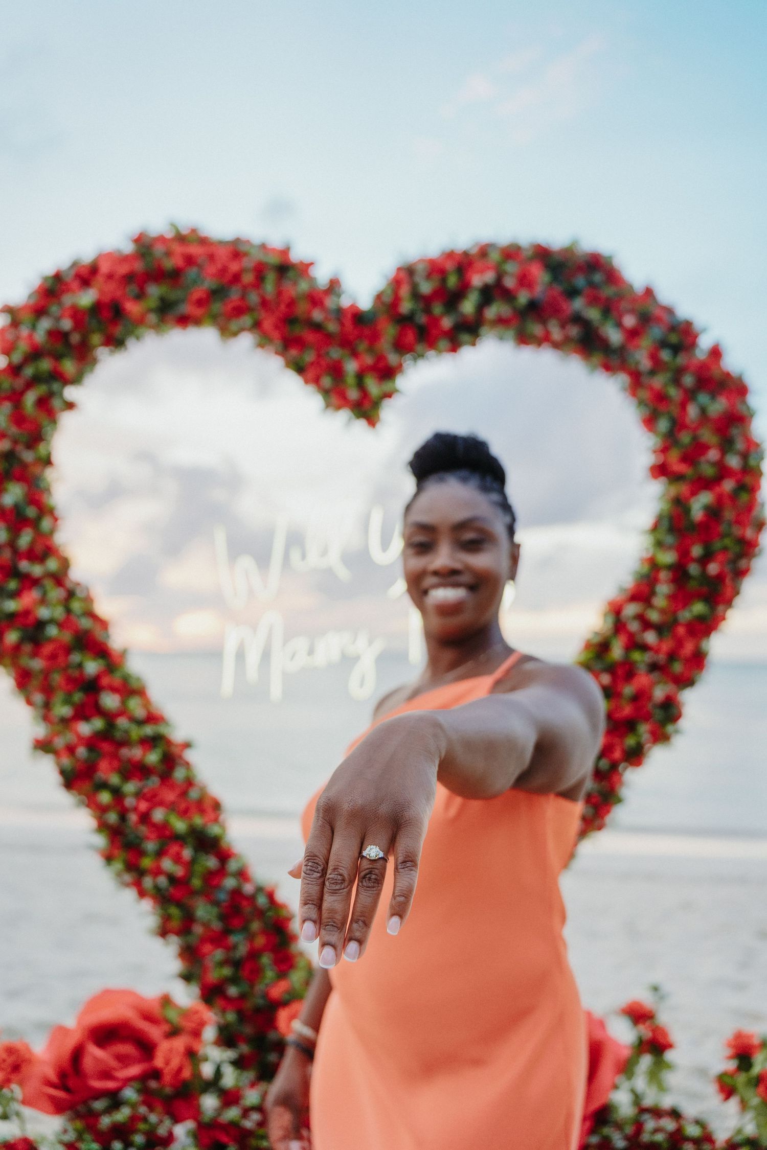 Smiling woman in orange dress shows off engagement ring in front of red rose heart arch.