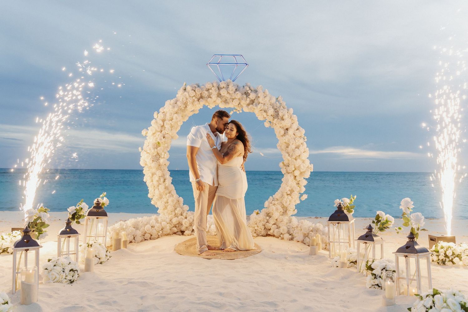 Couple embracing under a floral wedding arch on a tropical beach at sunset, surrounded by lanterns, flowers and sparkling fountain fireworks