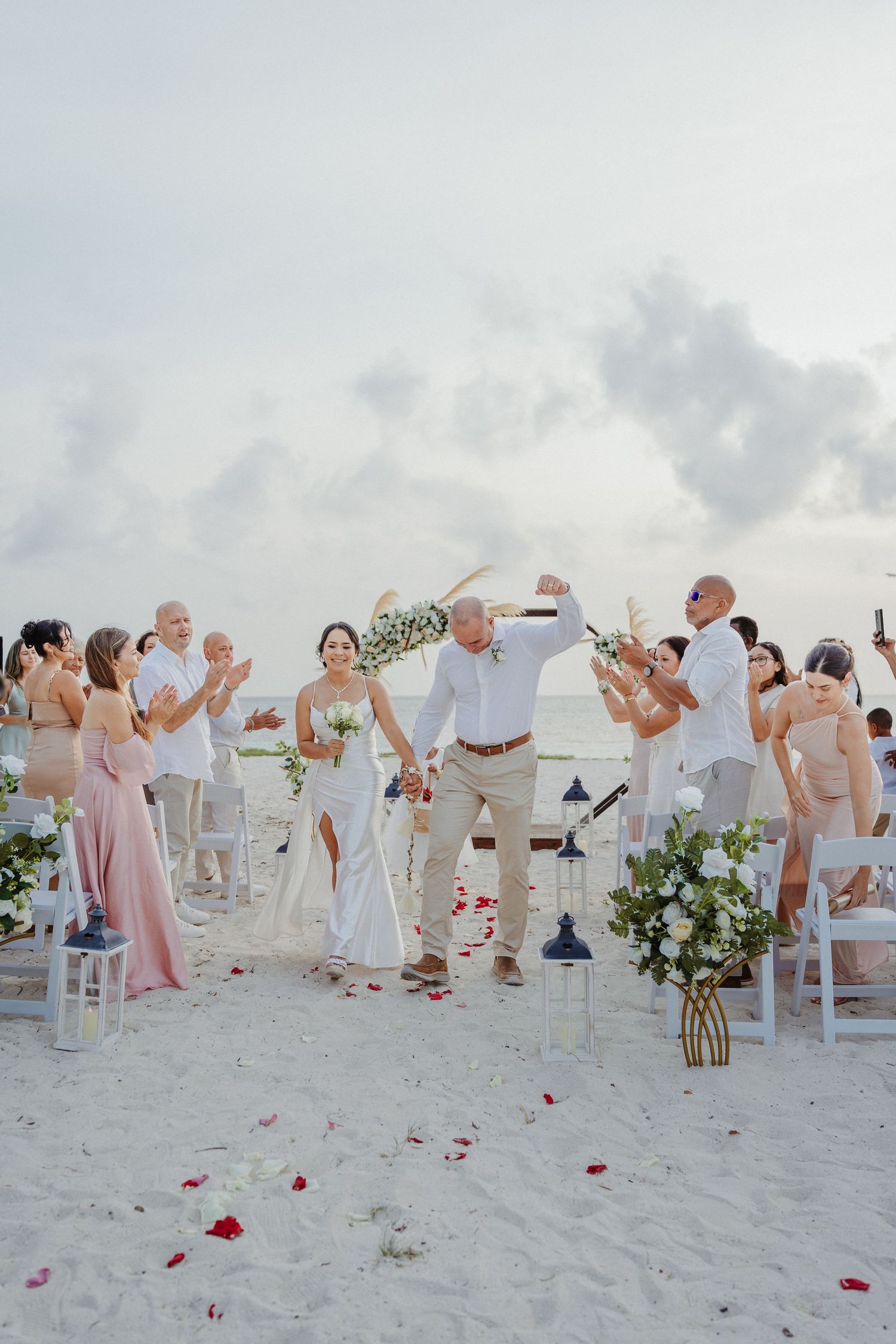 Newlyweds celebrating joyfully with guests cheering along flower petal-lined aisle at beach wedding ceremony.