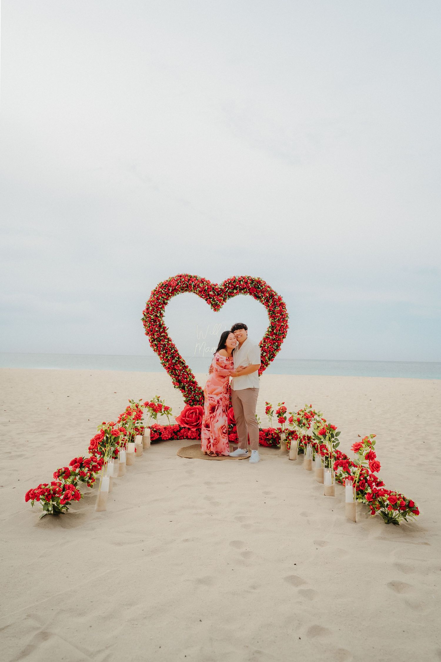 Couple hugging in front of a large red floral heart arch on a white sand beach.
