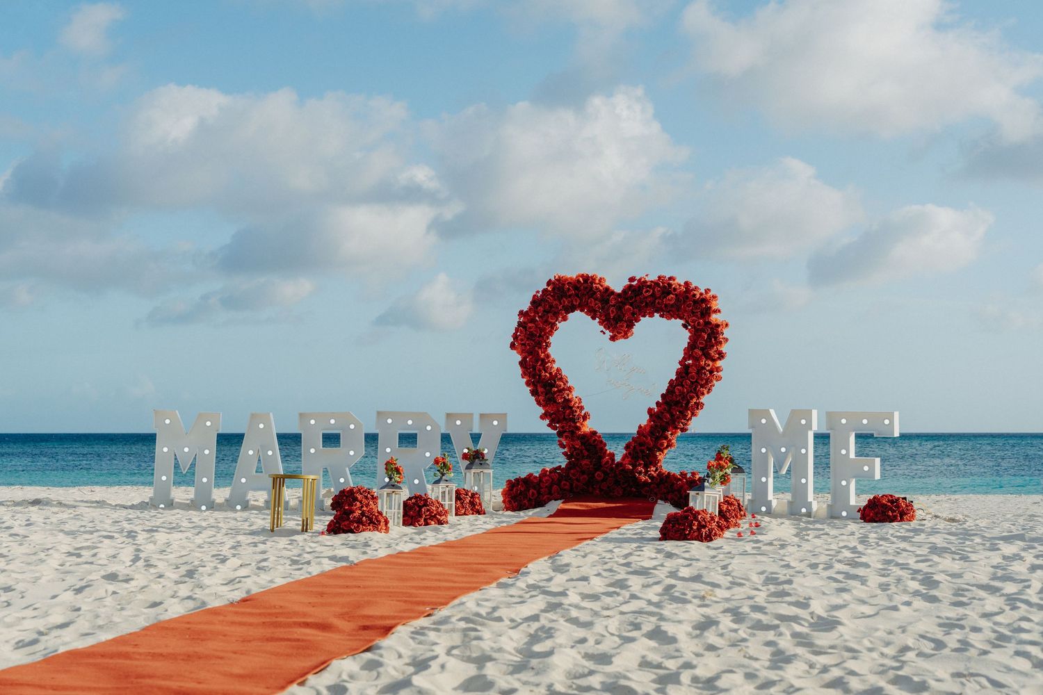 Beach wedding proposal setup with a red rose heart, orange aisle runner and white marquee letters on white sand beside the turquoise ocean.