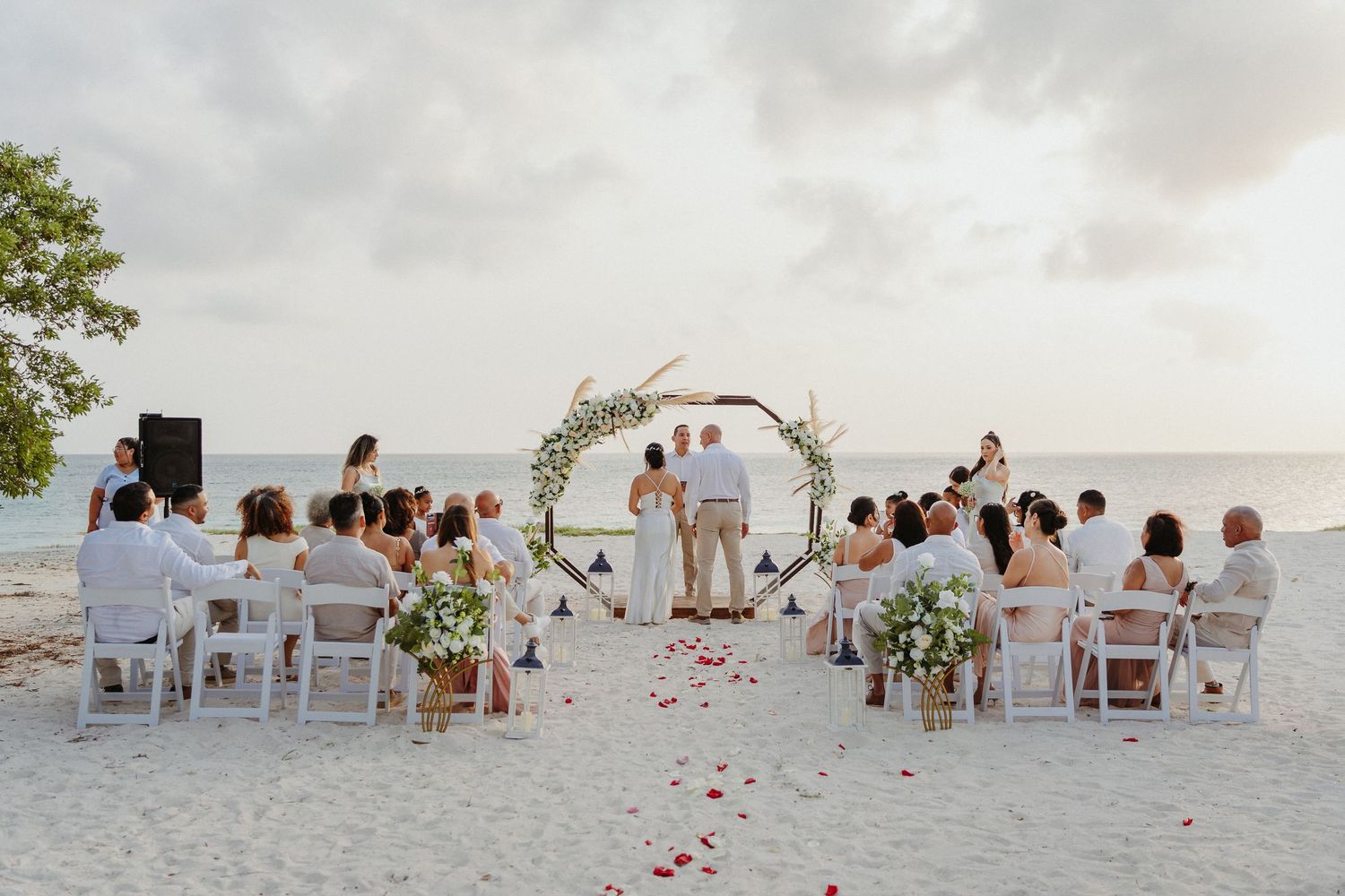 Beach wedding ceremony with guests seated on white chairs around a circular floral arch, rose petals on sand at sunset.