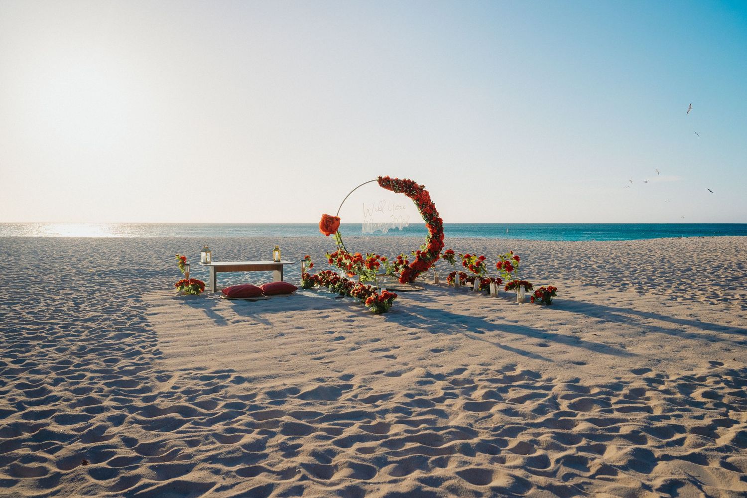 Romantic beach proposal setup with circular floral arch, flowers, and candles on white sandy beach.