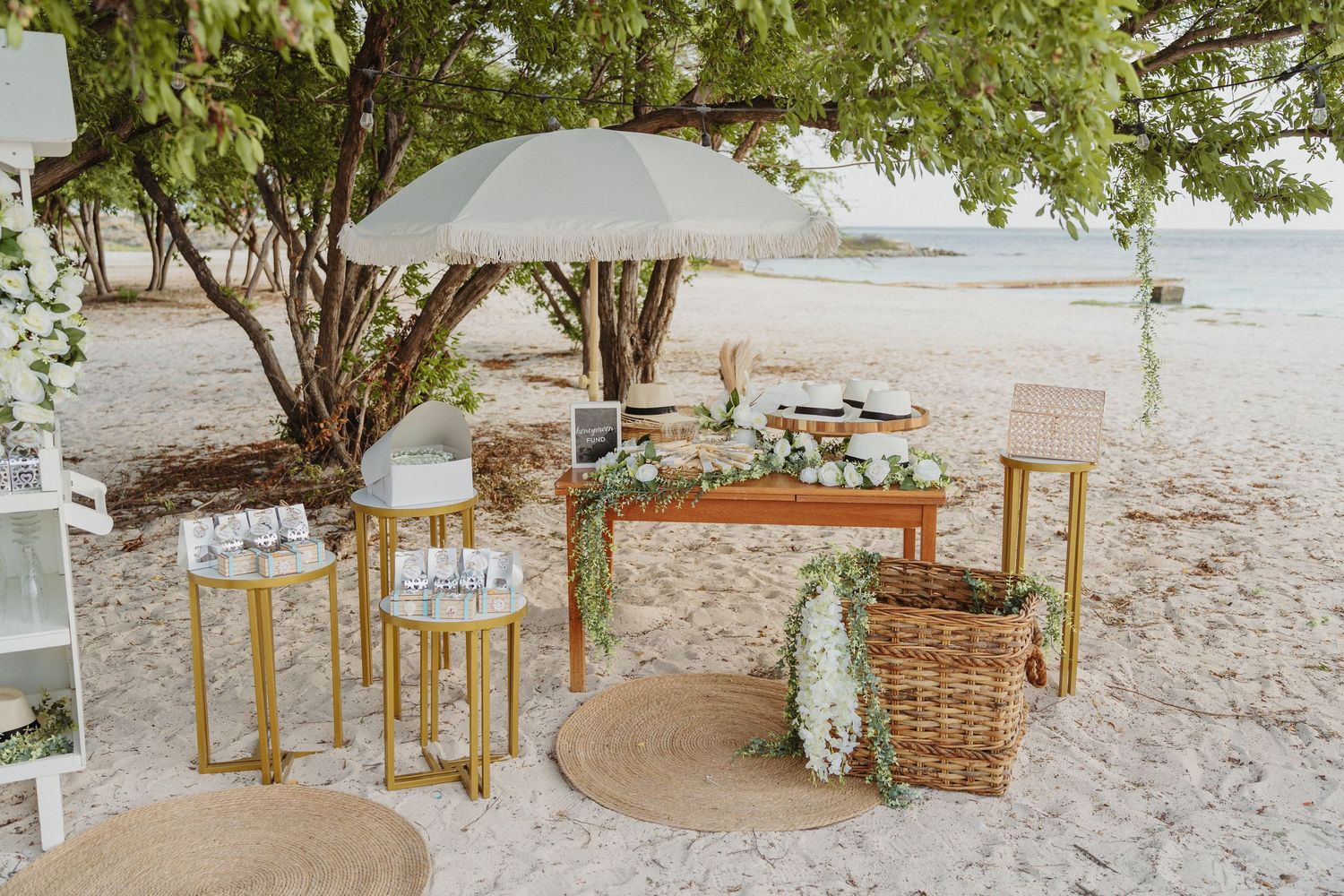 Elegant beach wedding setup with wooden tables, greenery, wicker basket, and white umbrella on white sand near the ocean.