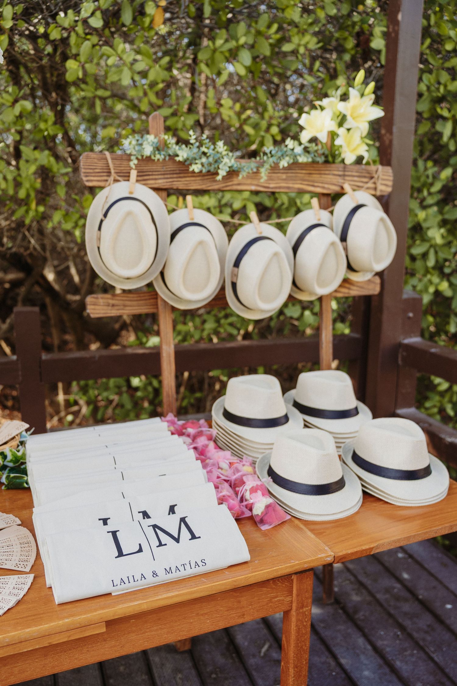 Wedding favor display with monogrammed LM tote bags and white Panama hats with navy bands on a wooden rack outdoors.