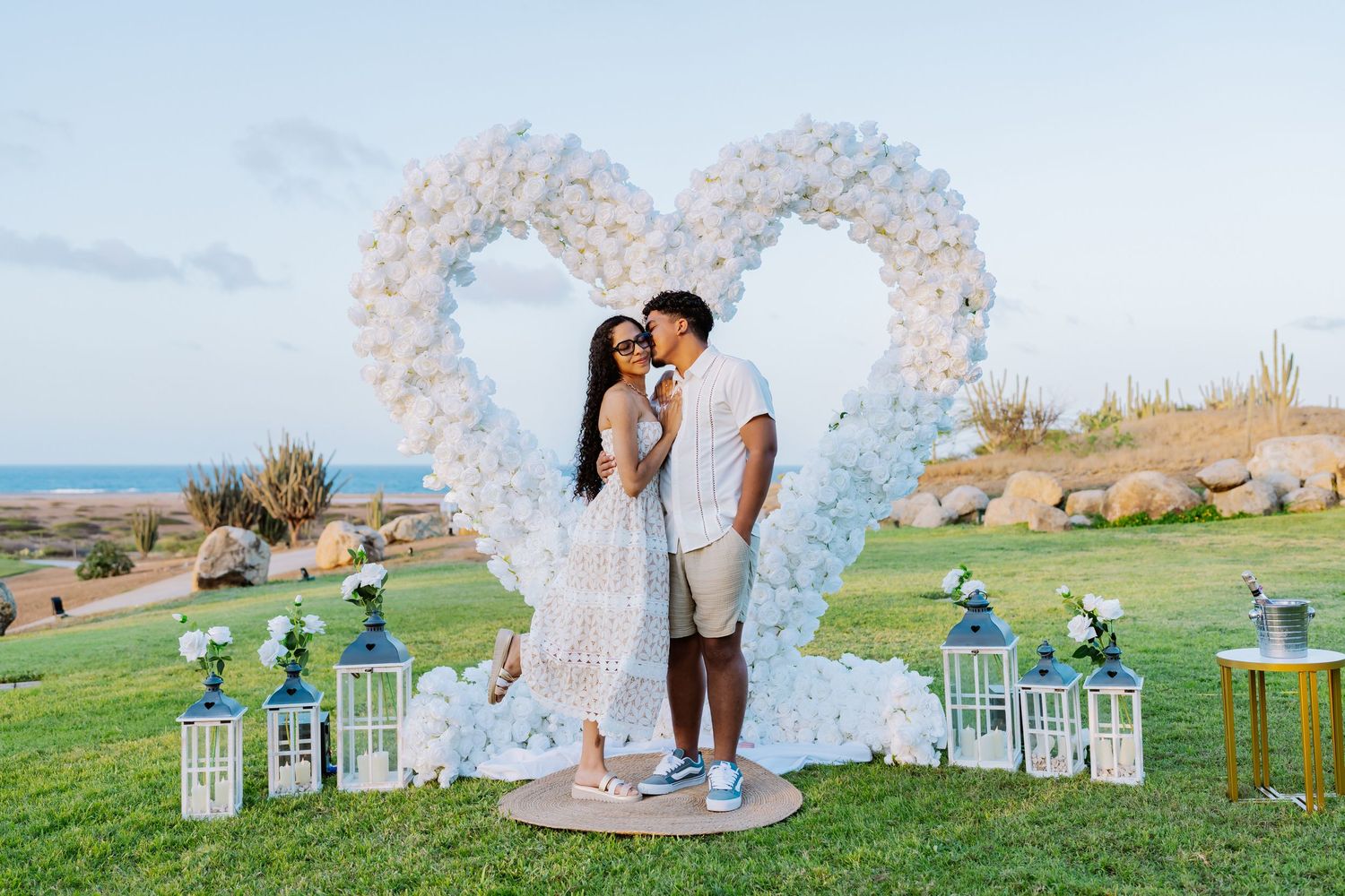 Couple sharing a romantic moment in front of a large white balloon heart arch at a seaside proposal setup.