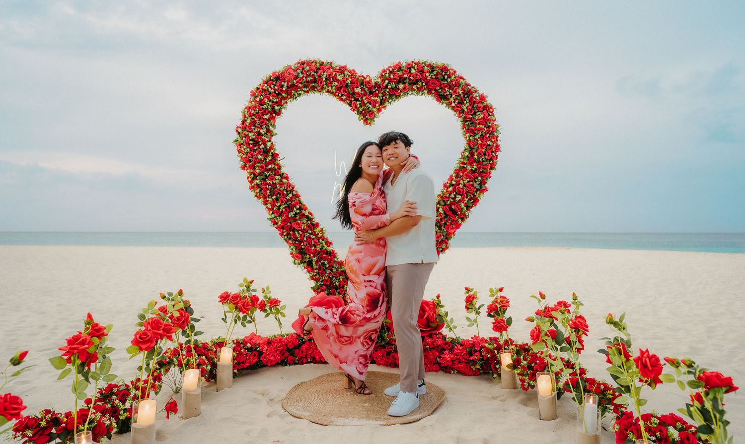 Couple embracing and smiling in front of a red rose heart arch with candles on the beach.