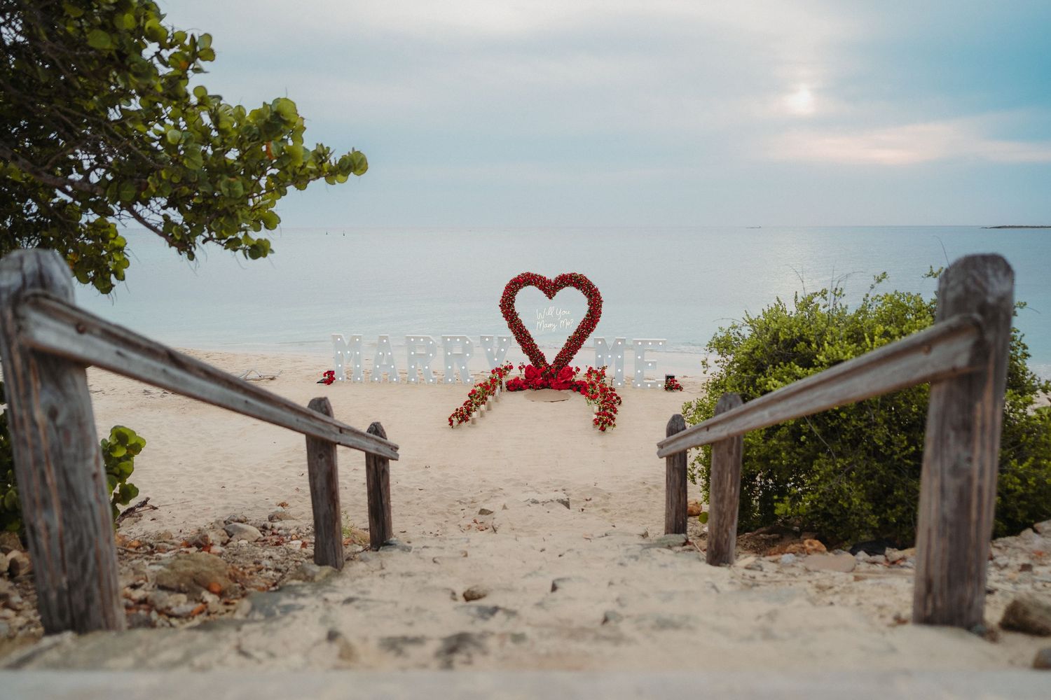 Red heart-shaped floral arch framed by wooden railings on a serene sandy beach.