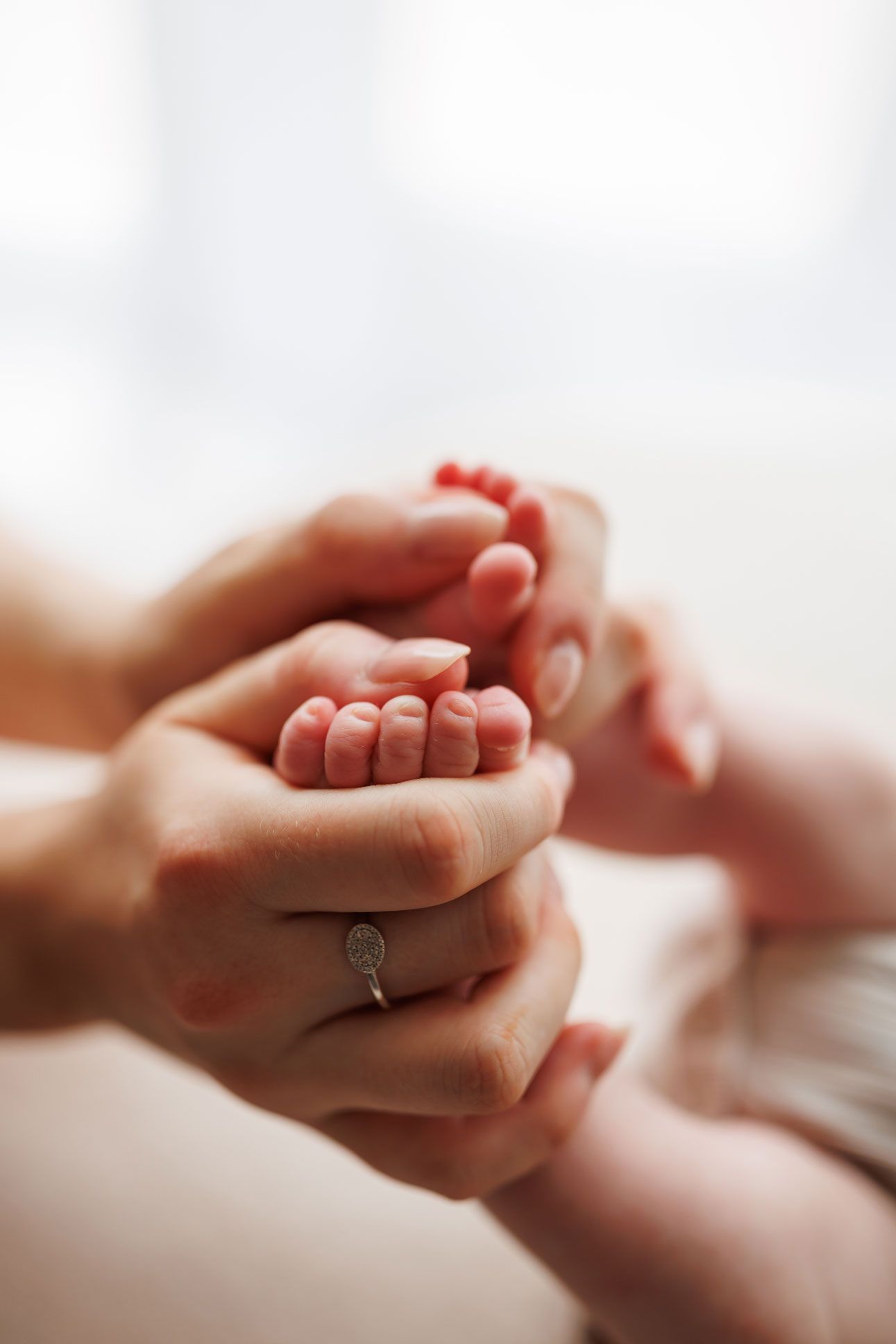 Adult hands gently cradling tiny newborn baby feet, with a diamond ring visible, symbolizing family love and new parenthood.