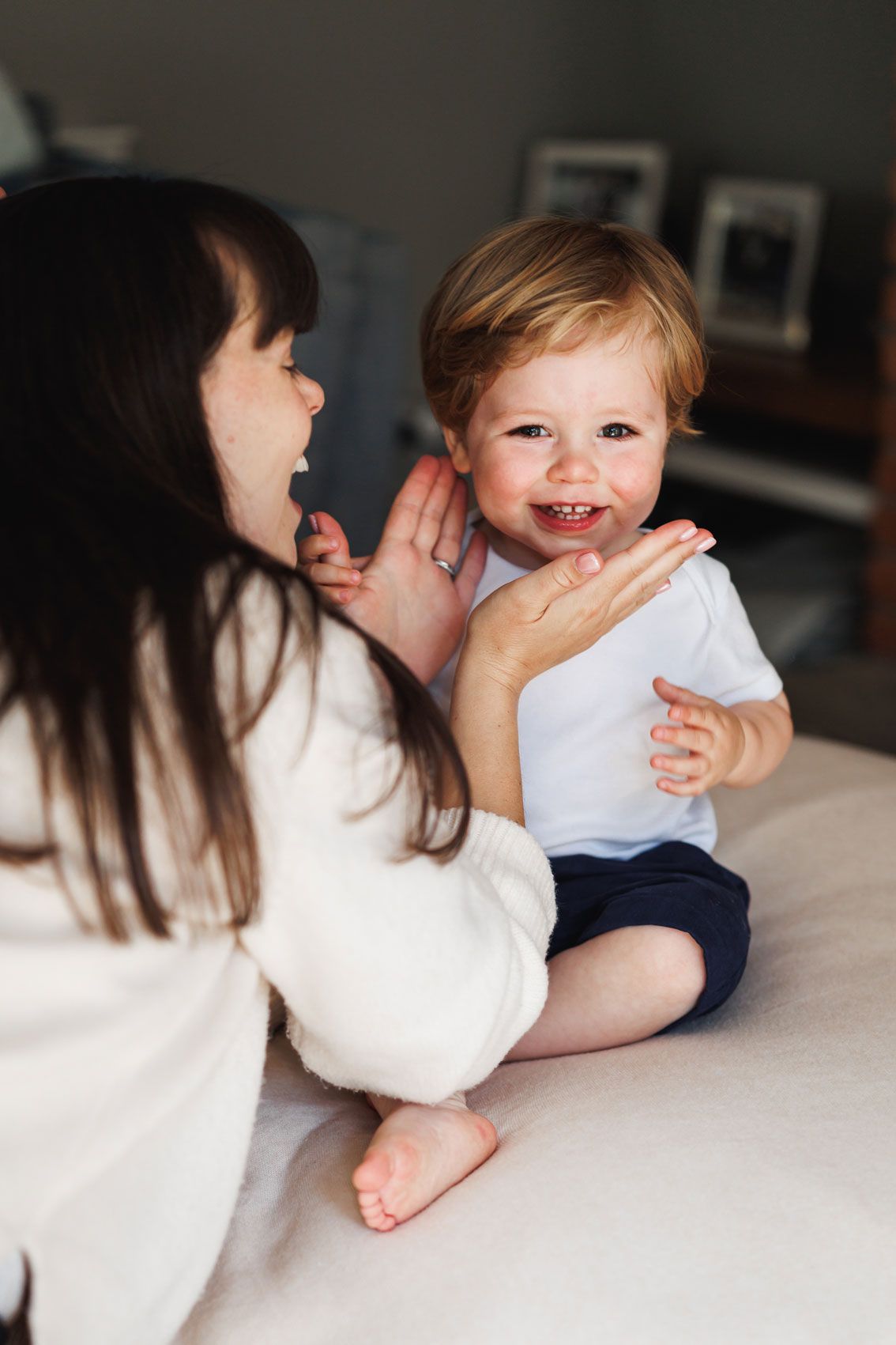 A laughing toddler in white shirt plays with dark-haired mother on bed during joyful indoor family moment.