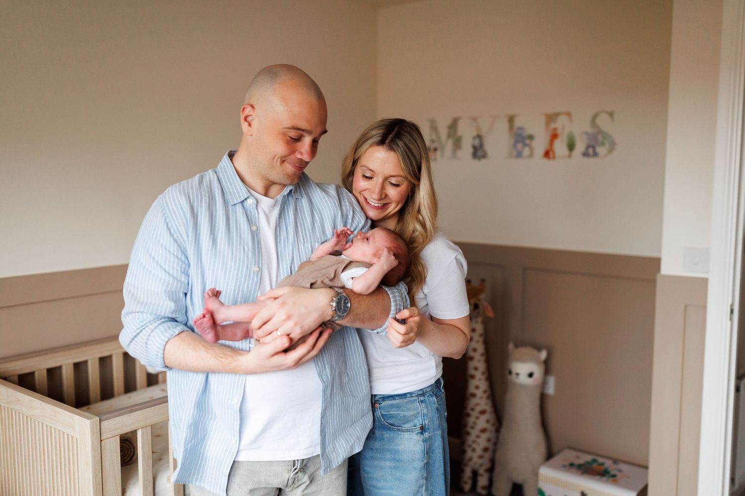 Parents standing in nursery room holding their newborn baby near a crib with name Myles.