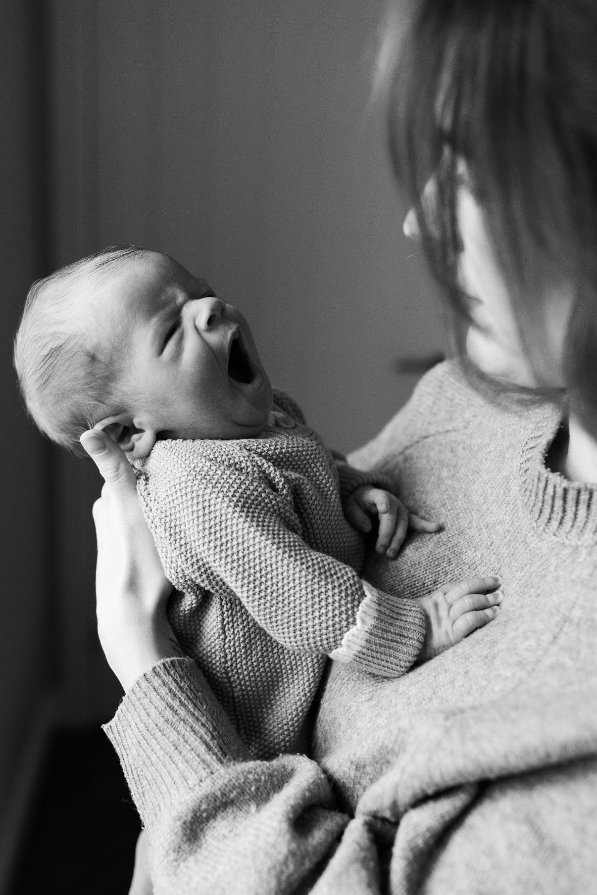 Black and white photo of a mother holding a yawning newborn baby in a knit outfit.