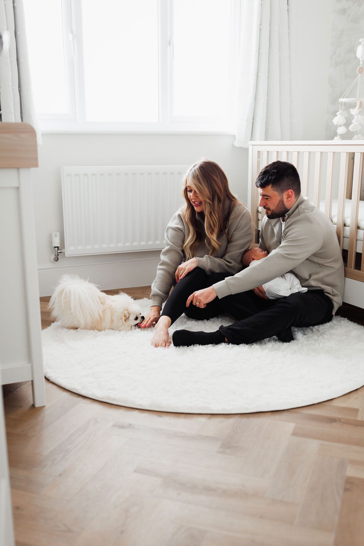Young couple sitting on a round white rug on hardwood floor playing with their fluffy white Pomeranian dog.