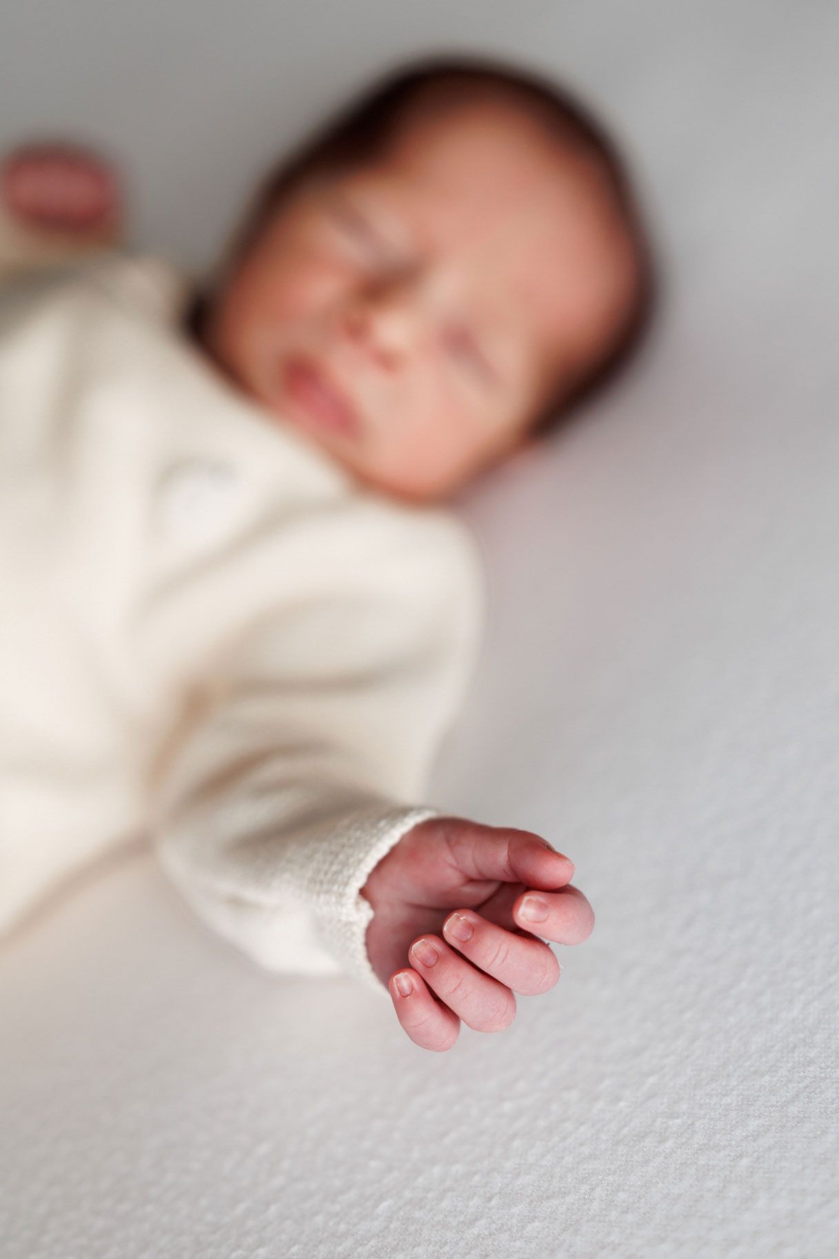 Sleeping newborn baby in a cream knit outfit lying on a soft white blanket with a tiny hand in sharp focus.