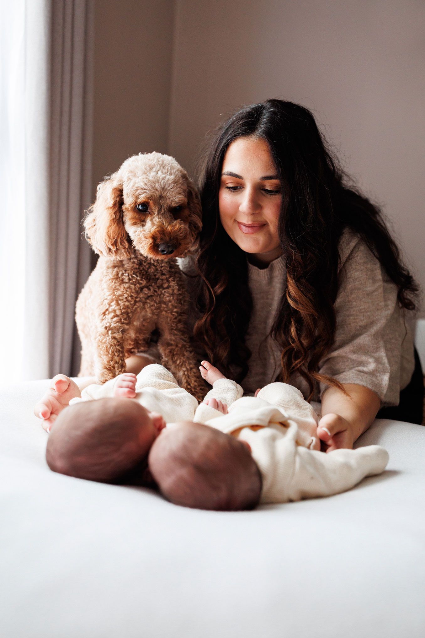 Smiling mother with long wavy hair sits on a bed beside a curious apricot poodle, gazing at two newborn twin babies in cream outfits.