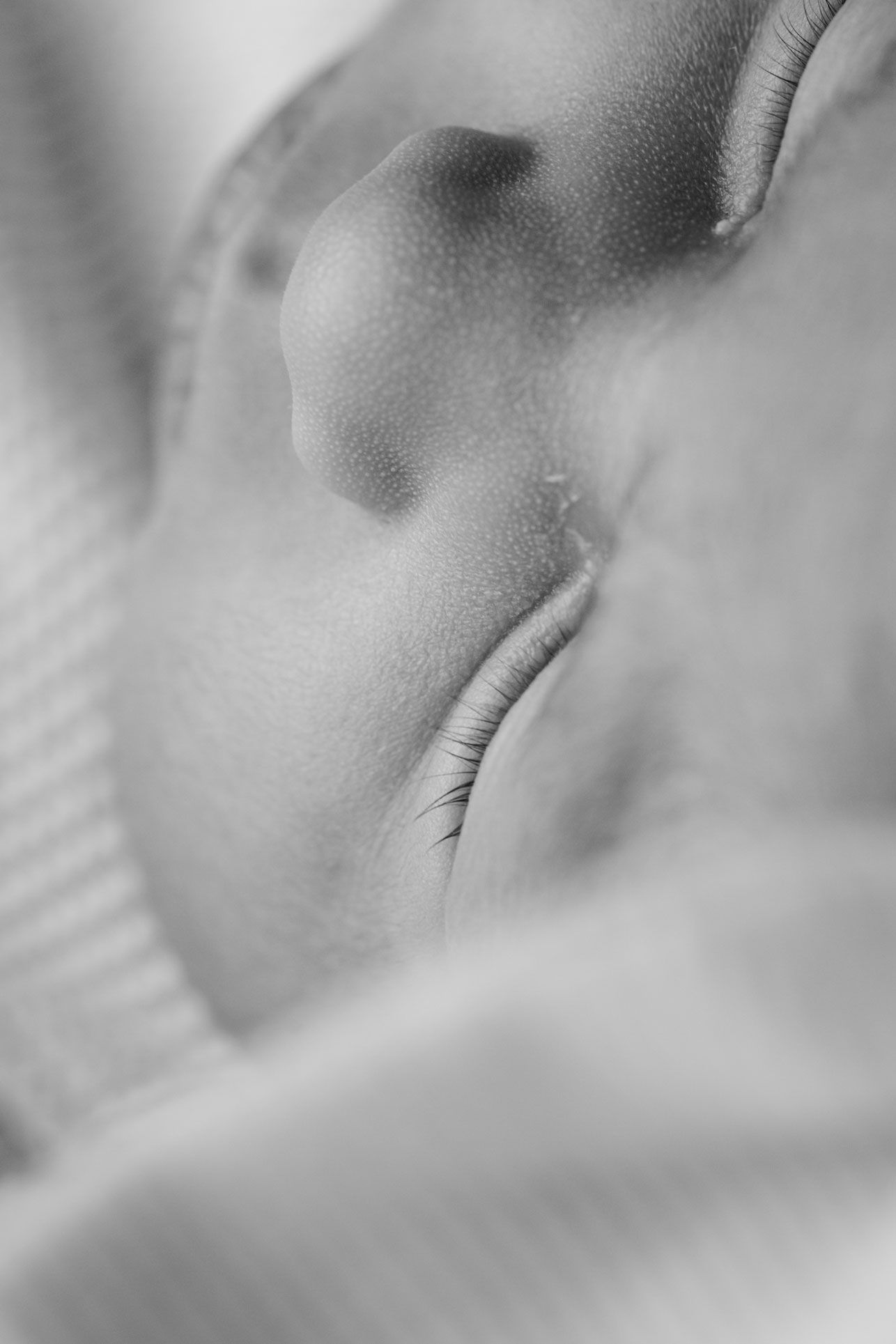 Black and white close-up of a sleeping newborn baby's nose and closed eye with delicate eyelashes, nestled in soft knit blankets.