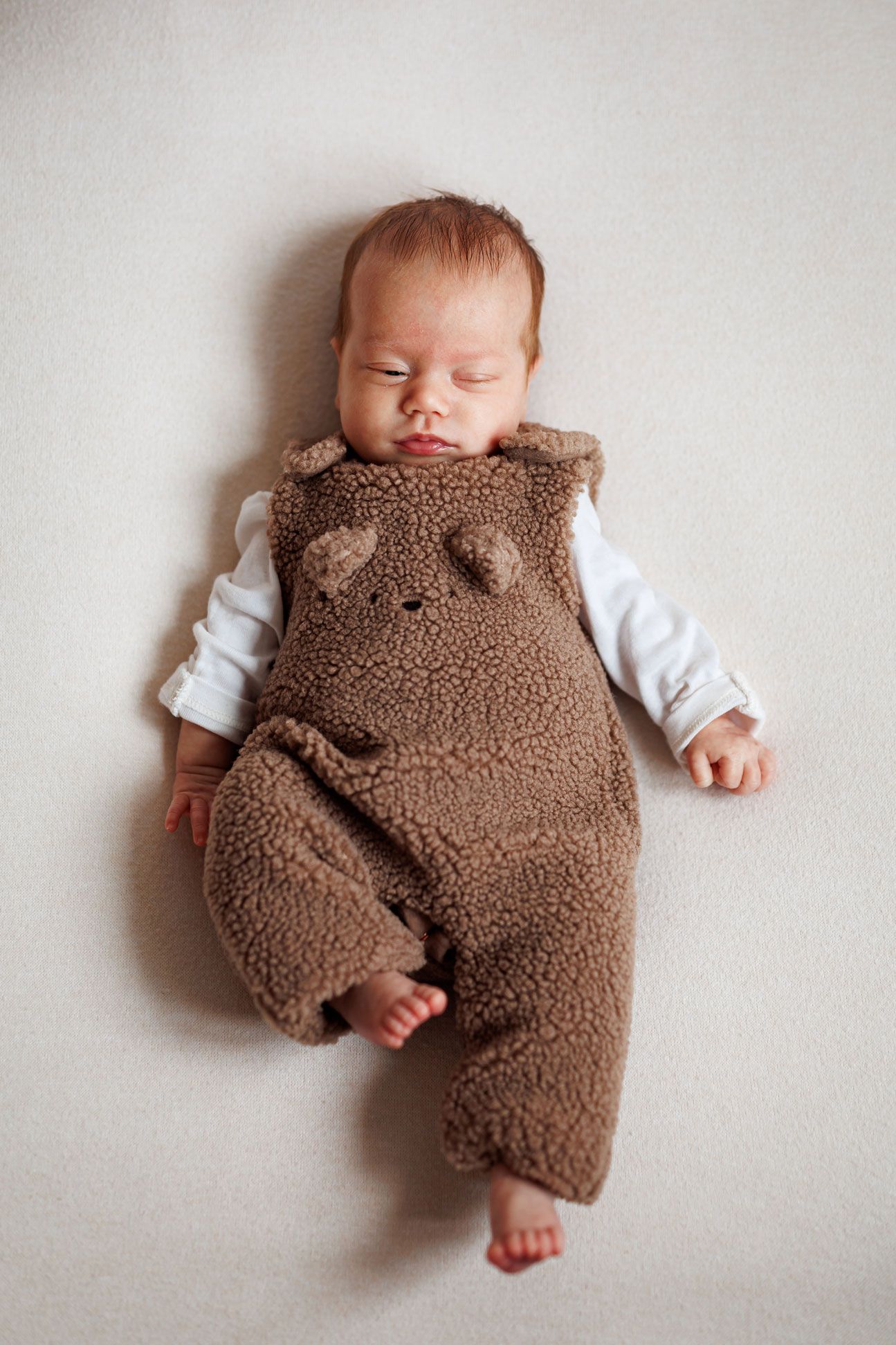 Sleeping newborn baby in a cozy brown teddy bear fleece romper with ears, lying on a soft cream blanket.