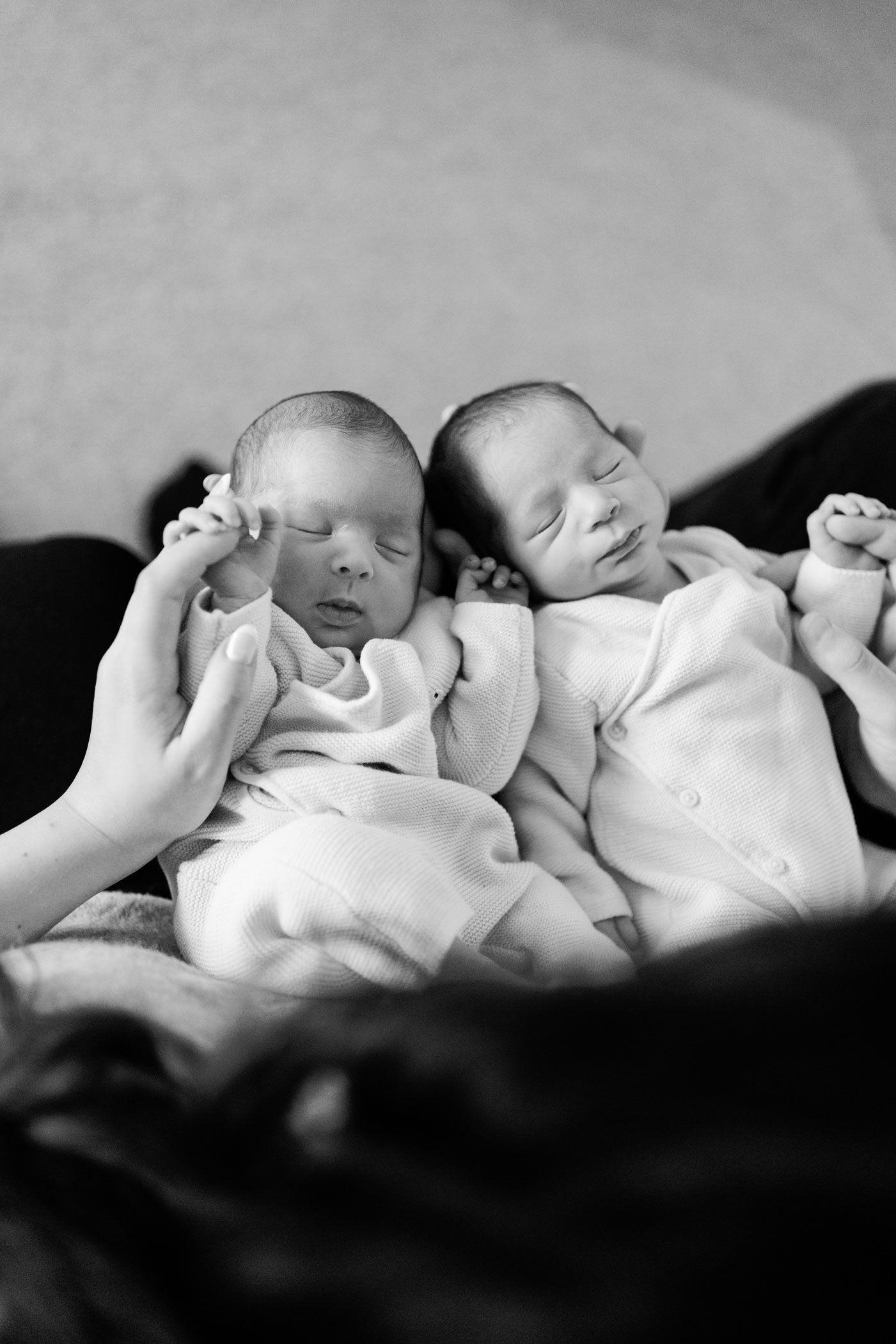 Black and white newborn photo of twin babies in soft white outfits sleeping side by side while a parent gently holds one tiny hand.