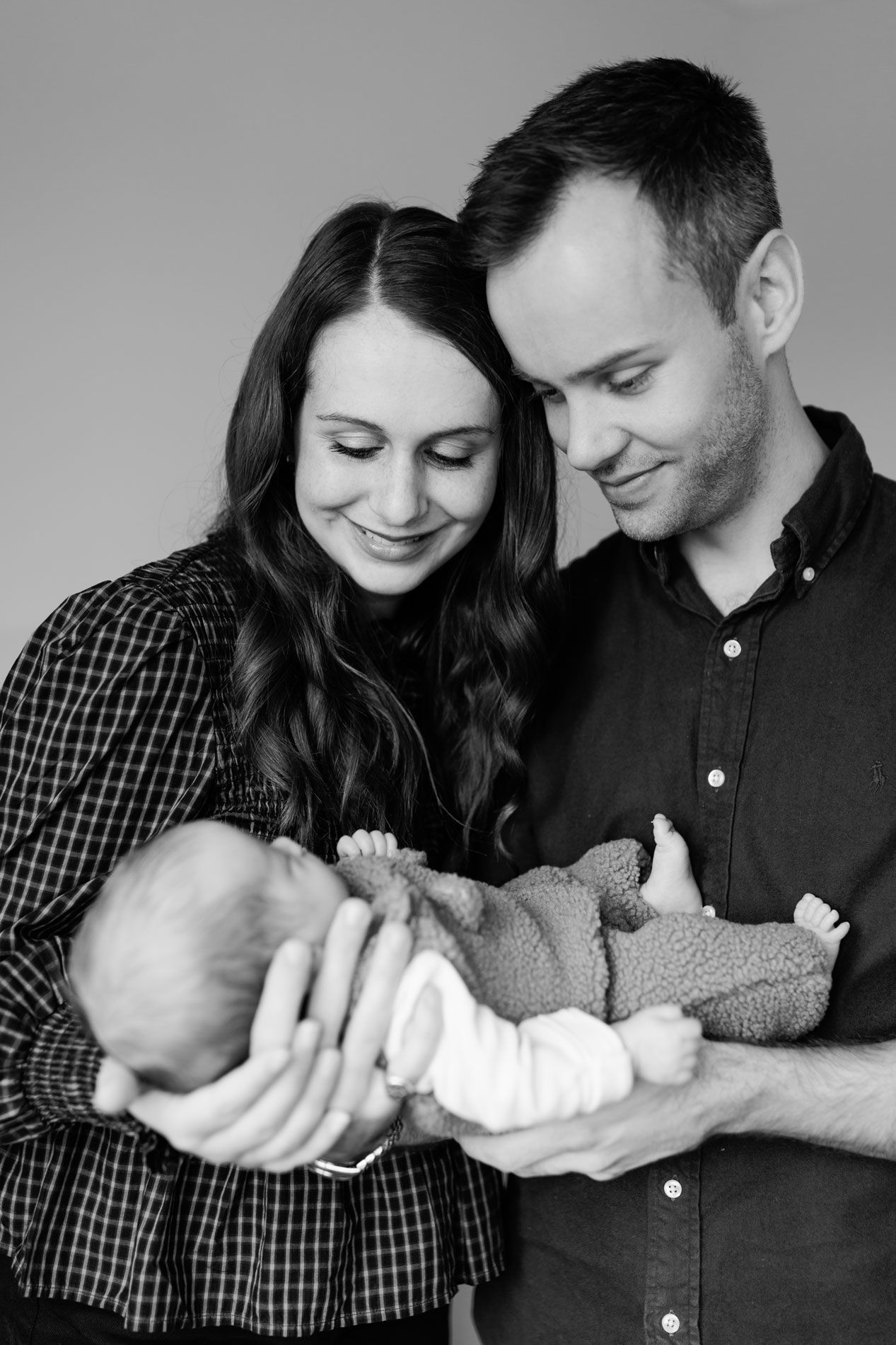 Black and white newborn portrait of smiling parents gently cradling their sleeping baby wrapped in a knit outfit.