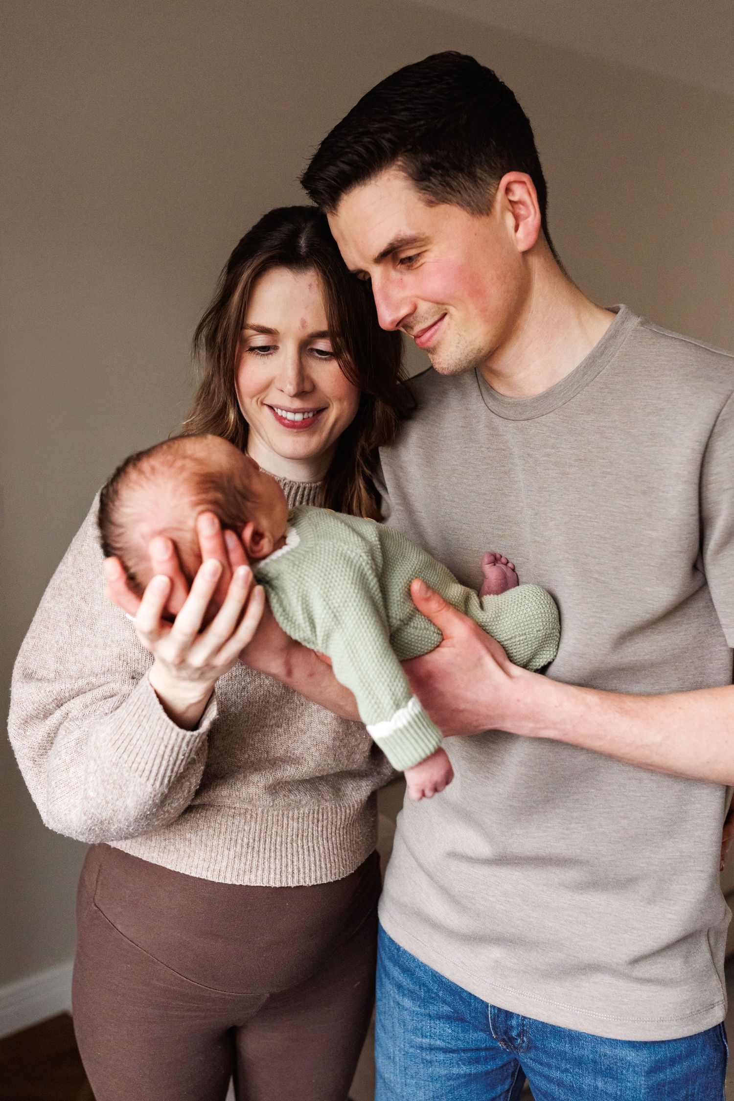 Smiling new parents in casual clothing hold their newborn baby dressed in a green knit outfit together.