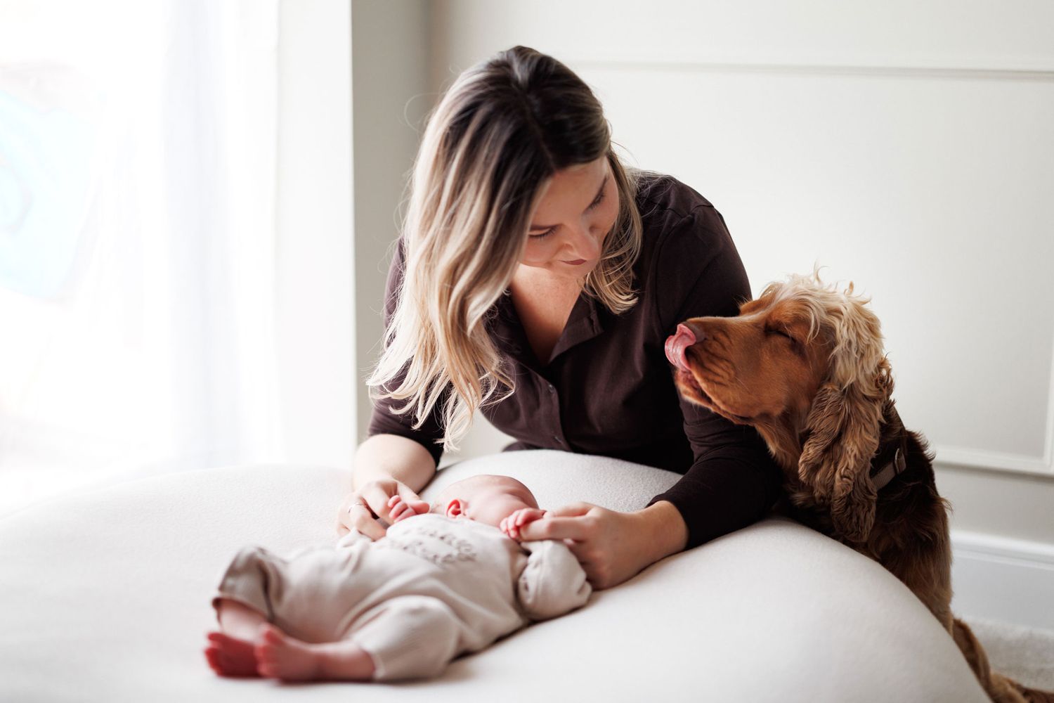 A mother with long blonde hair smiles down at her sleeping newborn baby while a golden cocker spaniel rests its head beside them on a bed.