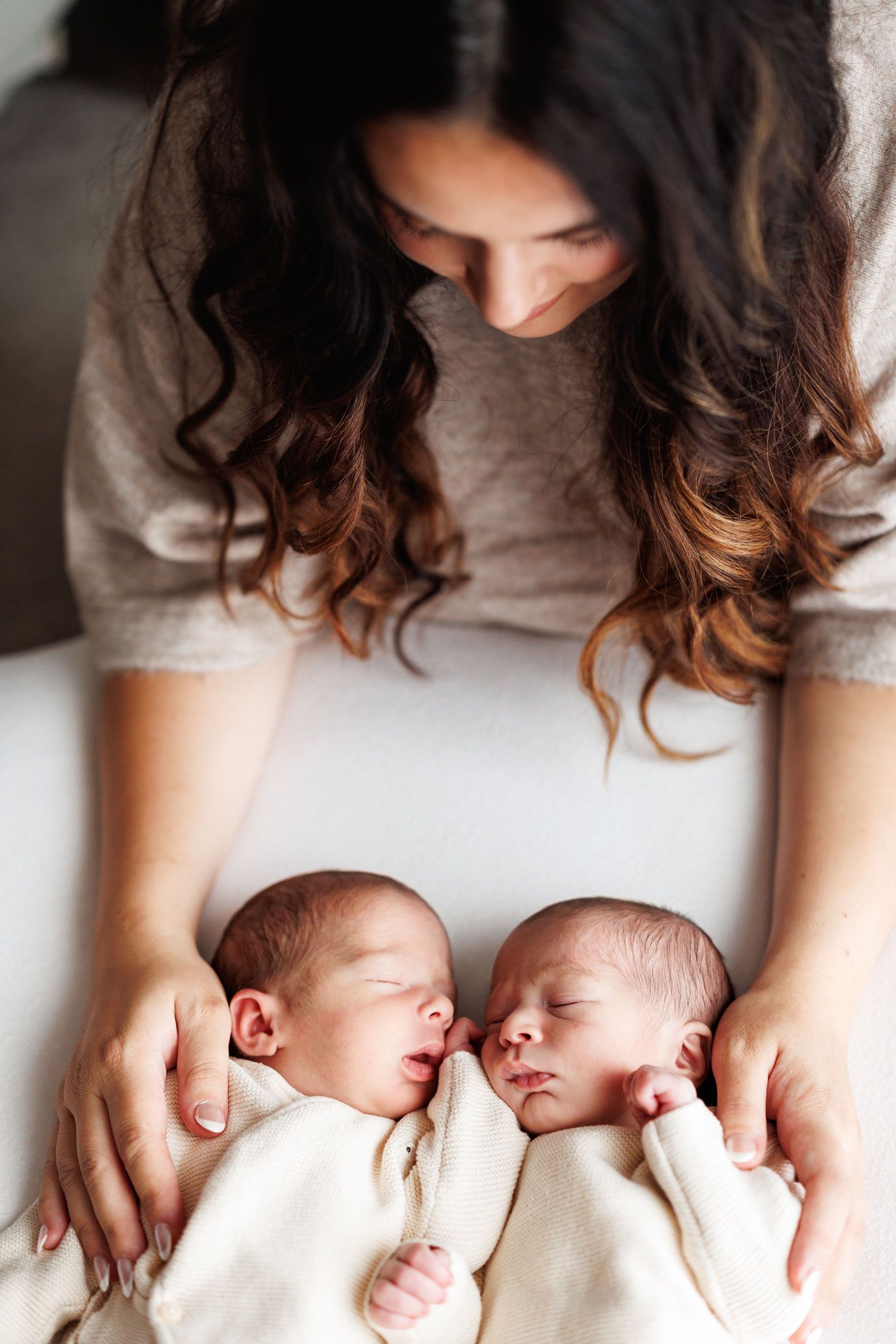Mother with long wavy hair gently cradles two sleeping newborn twins dressed in cream outfits, lying side by side in a soft newborn photo.