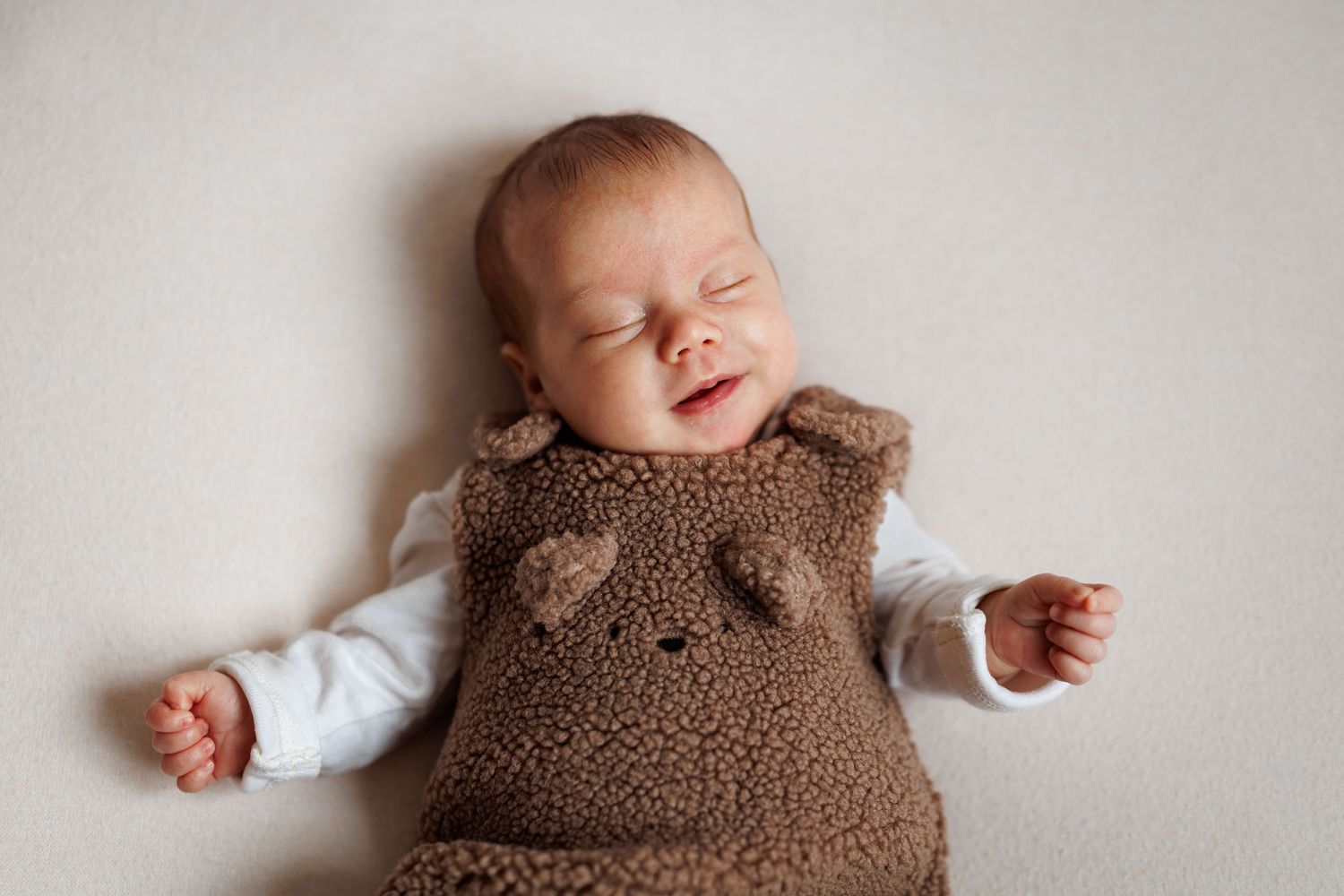 Sleeping newborn baby in a brown teddy bear fleece romper with ear details, arms resting at sides on a soft cream background.