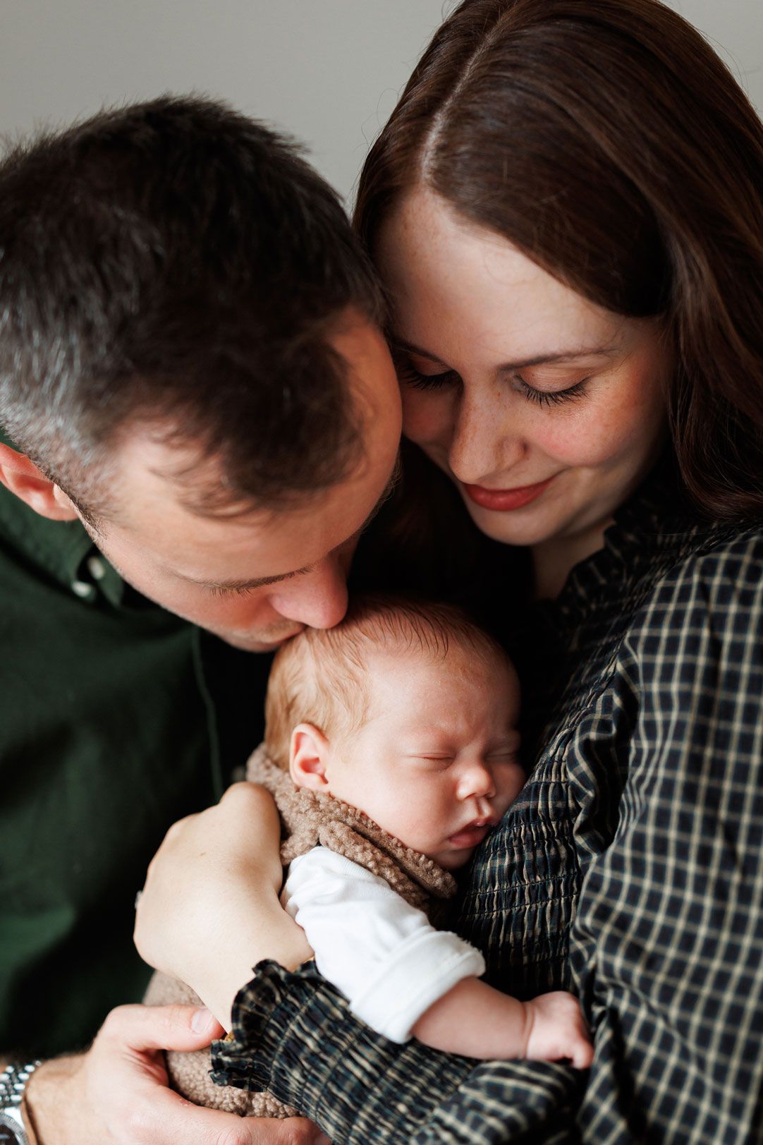 A father and mother tenderly embrace their sleeping newborn baby, with the dad kissing the infant's forehead in a warm family portrait.